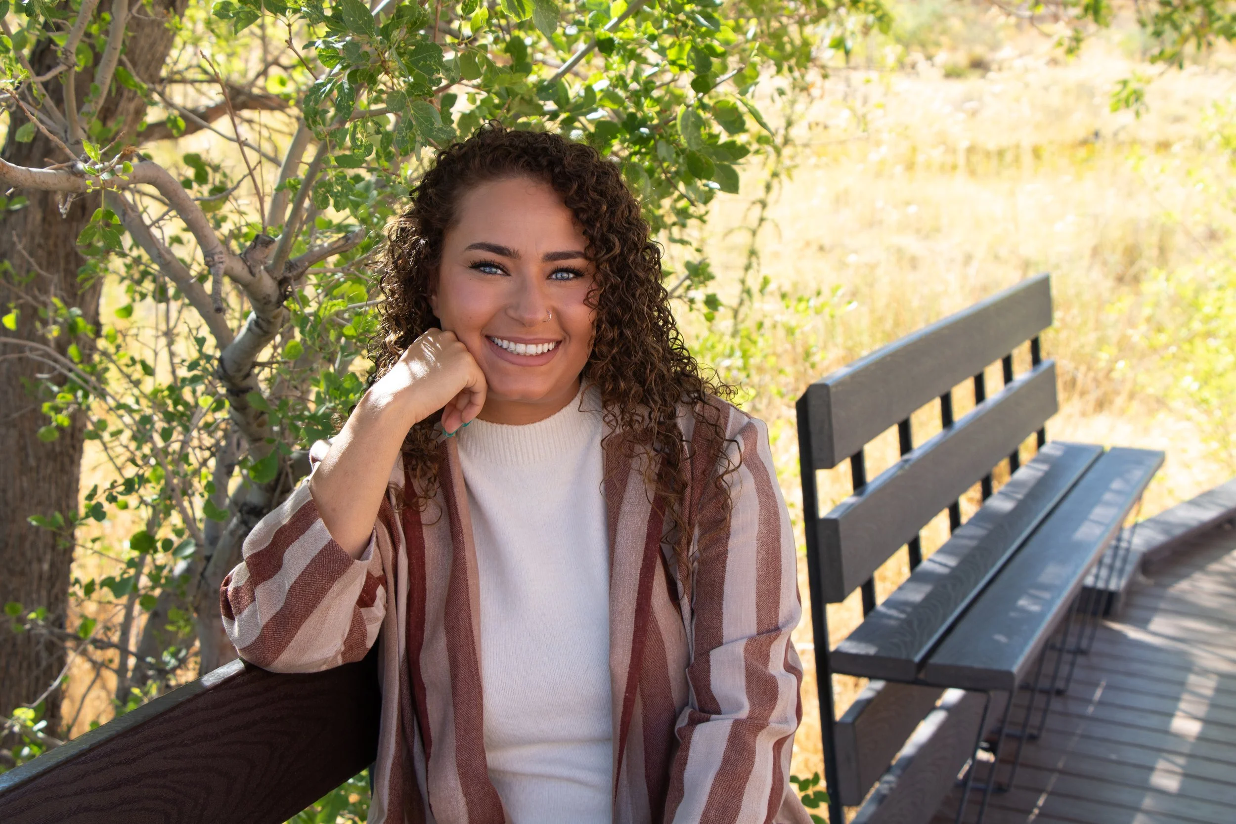 A woman with curly hair and blue eyes smiling while sitting on a park bench amidst green trees and autumn foliage.