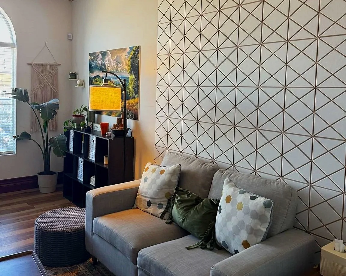 Living room with beige sofa, decorative pillows, patterned accent wall, large window, houseplants, and a black shelving unit with art and decor.