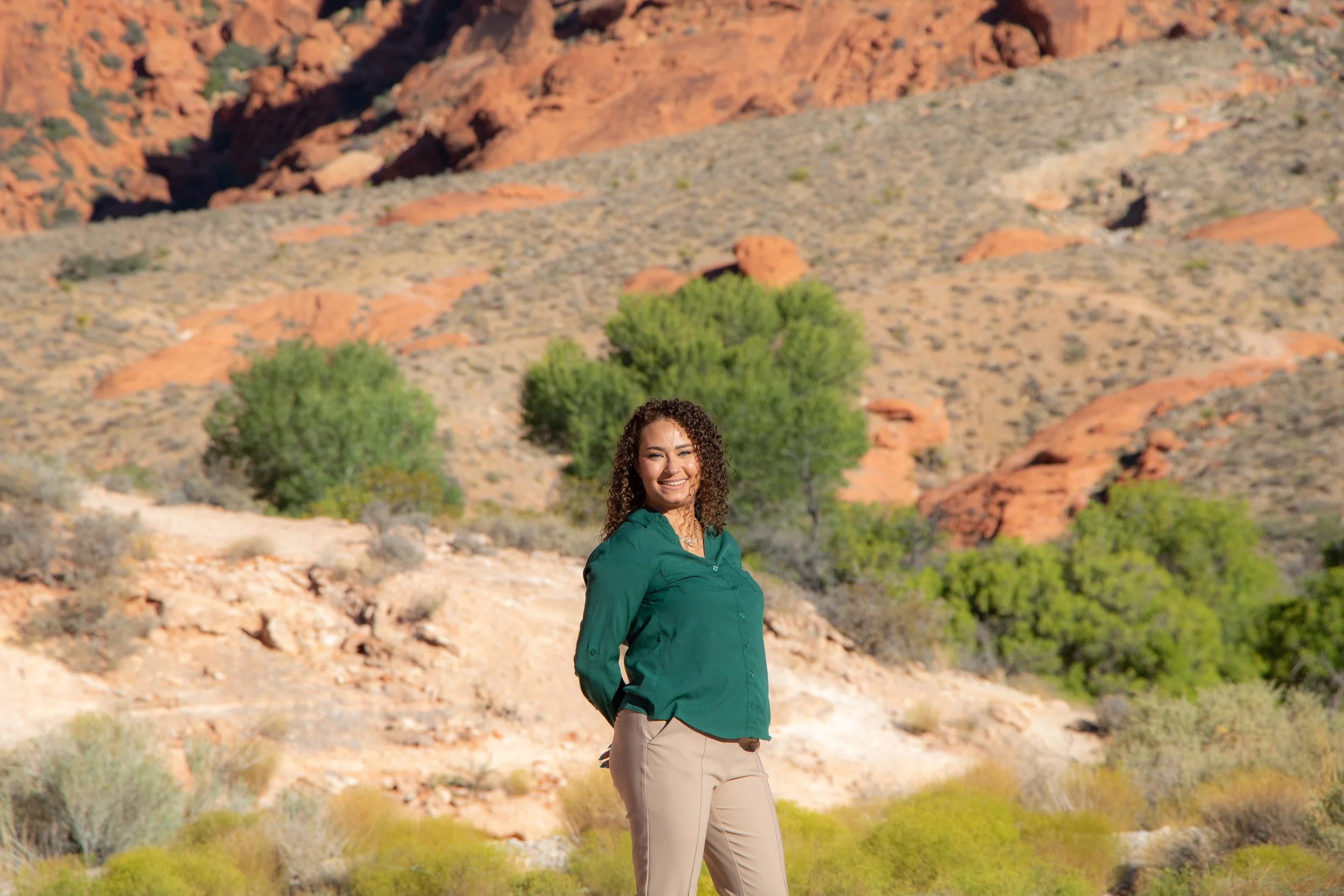 A woman with curly hair smiling outdoors in front of a desert landscape with red rock formations and sparse green trees.