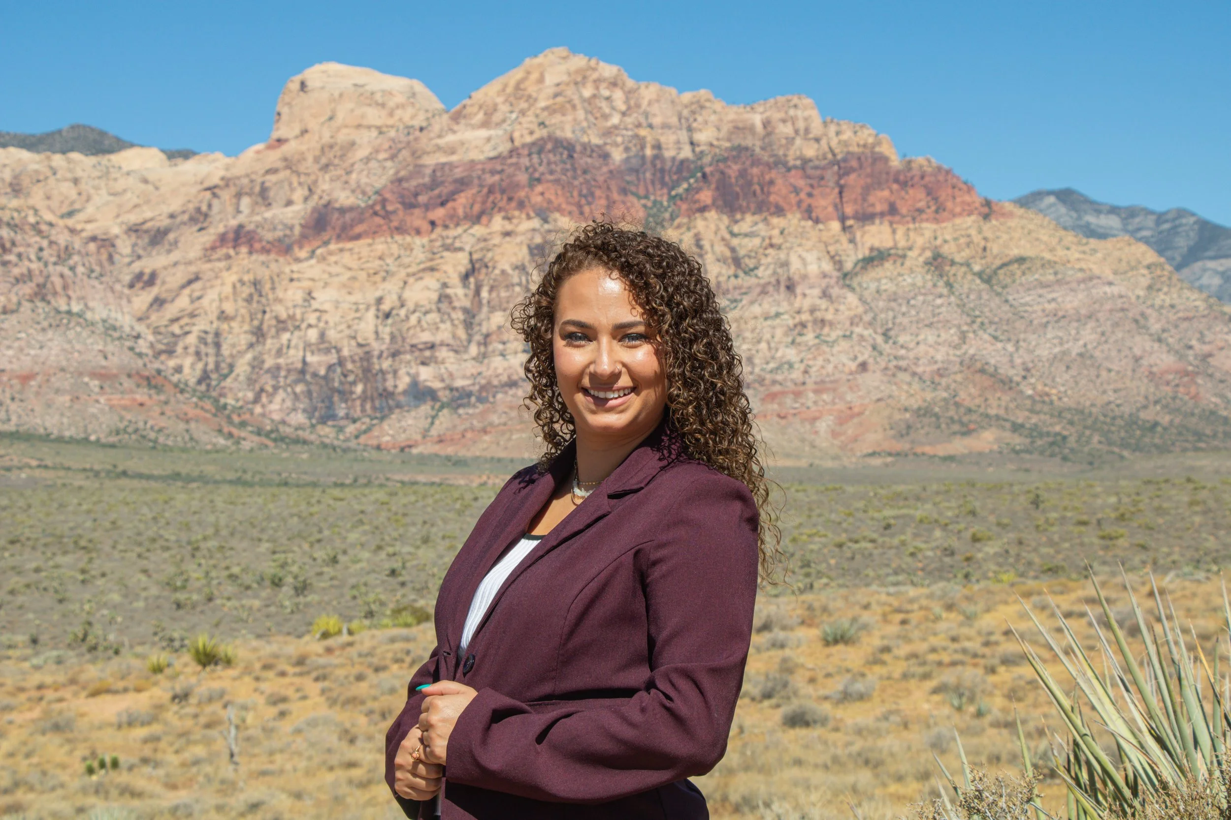 A woman with curly hair smiling outdoors in front of a desert mountain landscape with blue sky, wearing a maroon blazer and white top.