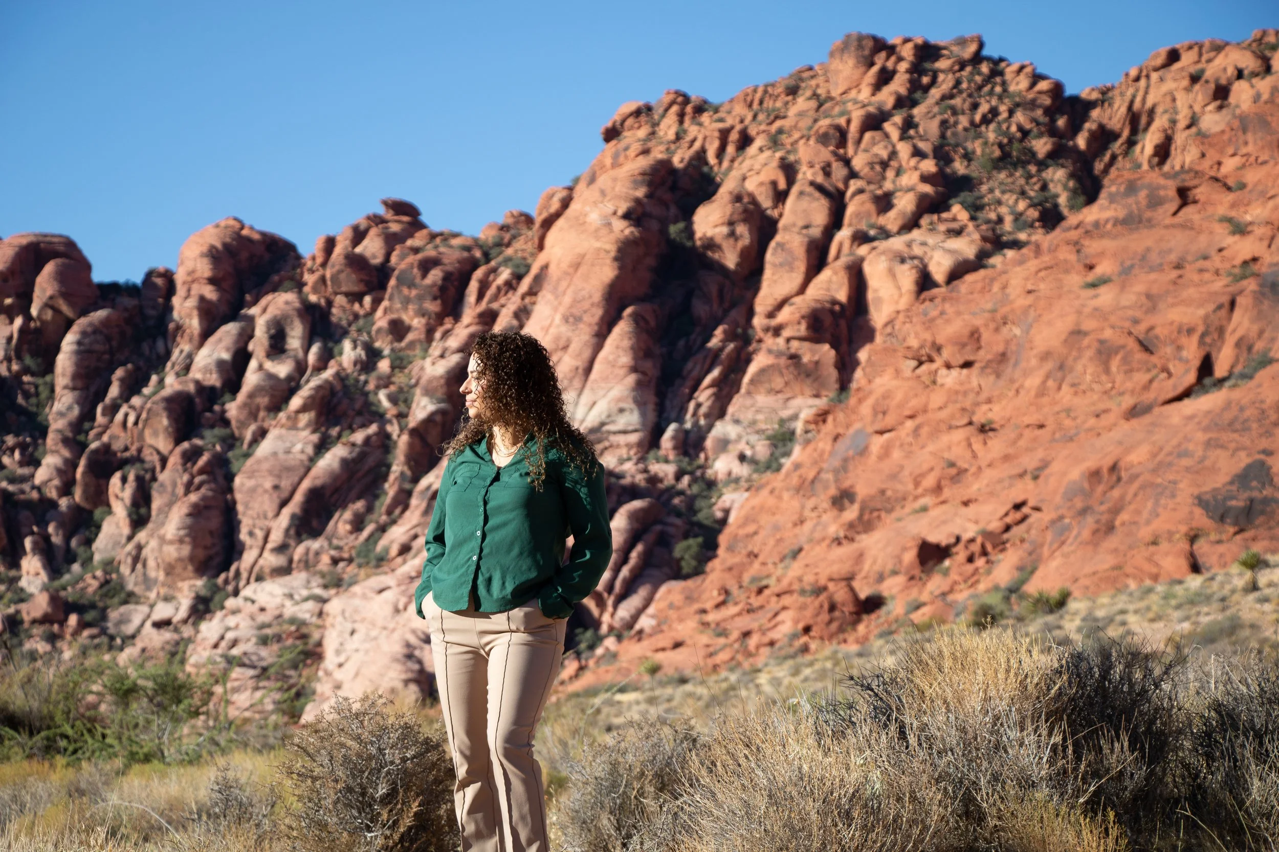 A woman with curly hair wearing a green jacket and beige pants standing outdoors near red rock formations under a clear blue sky.