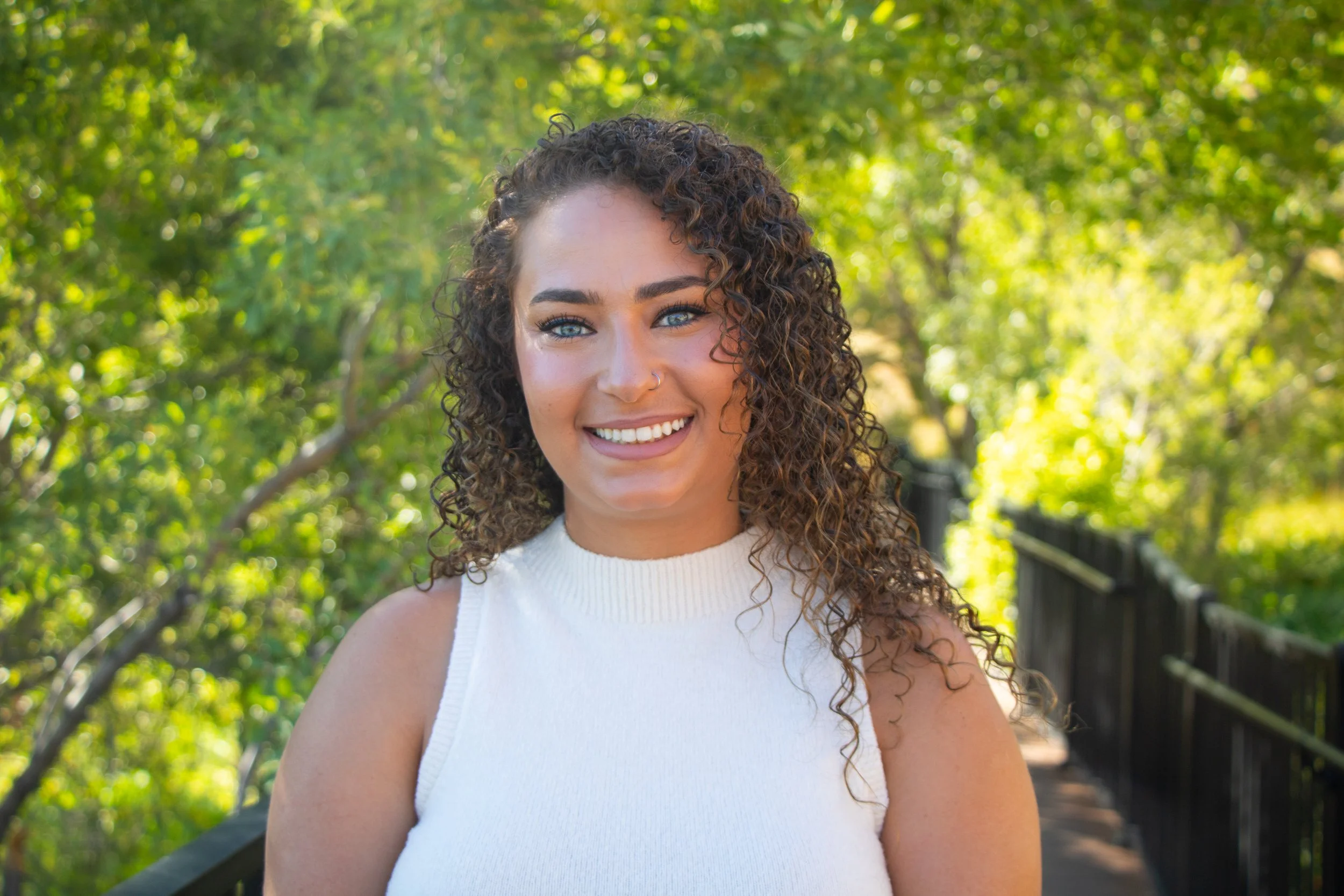 A young woman with curly hair, bright blue eyes, and a nose piercing, smiling outdoors on a sunny day with green trees and a black fence in the background.