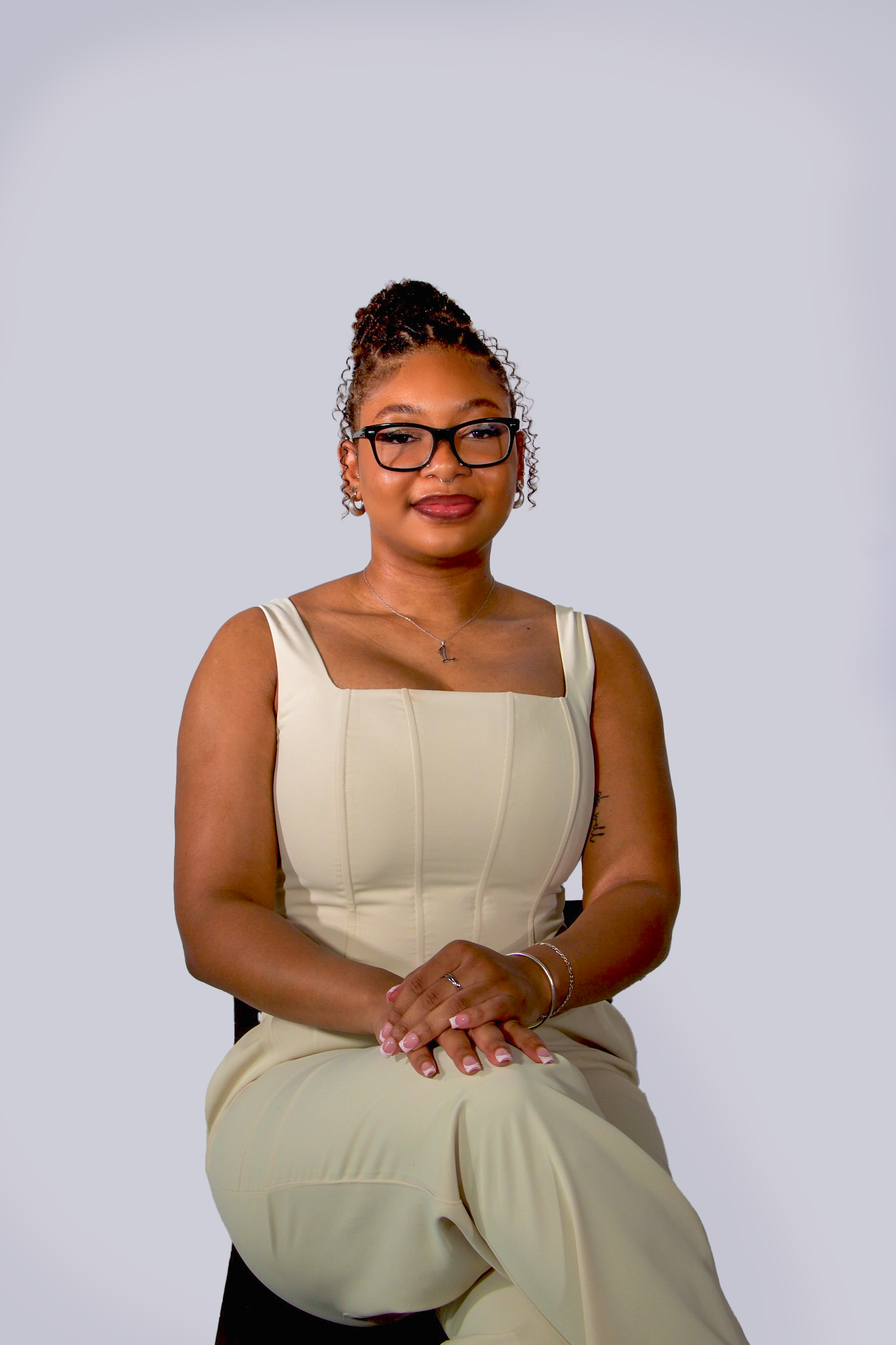 A graphic designer with glasses and curly hair styled in a high bun, wearing a cream-colored dress and jewelry, sitting with her hands clasped on her lap against a plain light gray background.