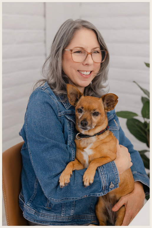 A woman with gray hair and glasses smiling while holding a small tan dog indoors near a white brick wall and some green plants.