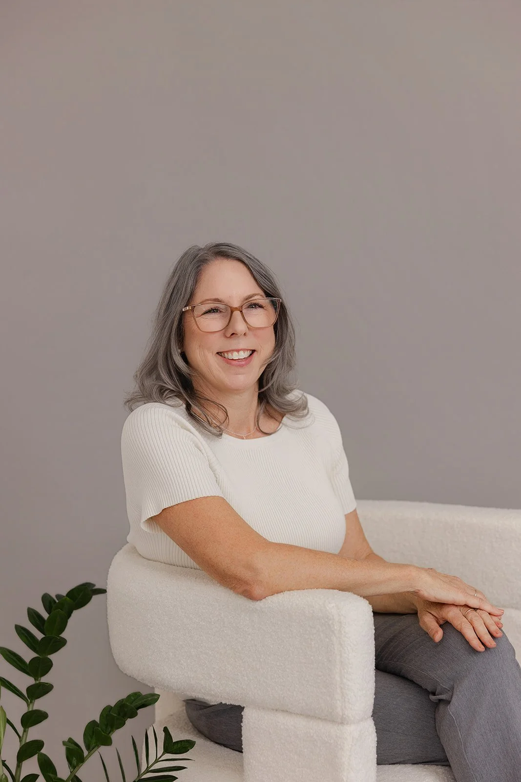 A smiling middle-aged woman with gray hair, wearing glasses, a white short-sleeve top, and gray pants, sitting on a white textured armchair against a plain light gray background with green houseplants nearby.