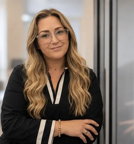 A woman with long wavy blonde hair, wearing glasses, a black blouse with white striped accents, and a bracelet on her wrist, standing indoors against a blurred background.