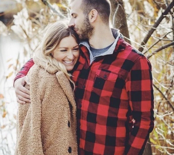 A couple sharing a warm hug outdoors in fall, surrounded by trees with autumn leaves. The woman smiles happily, and the man kisses her on the forehead.
