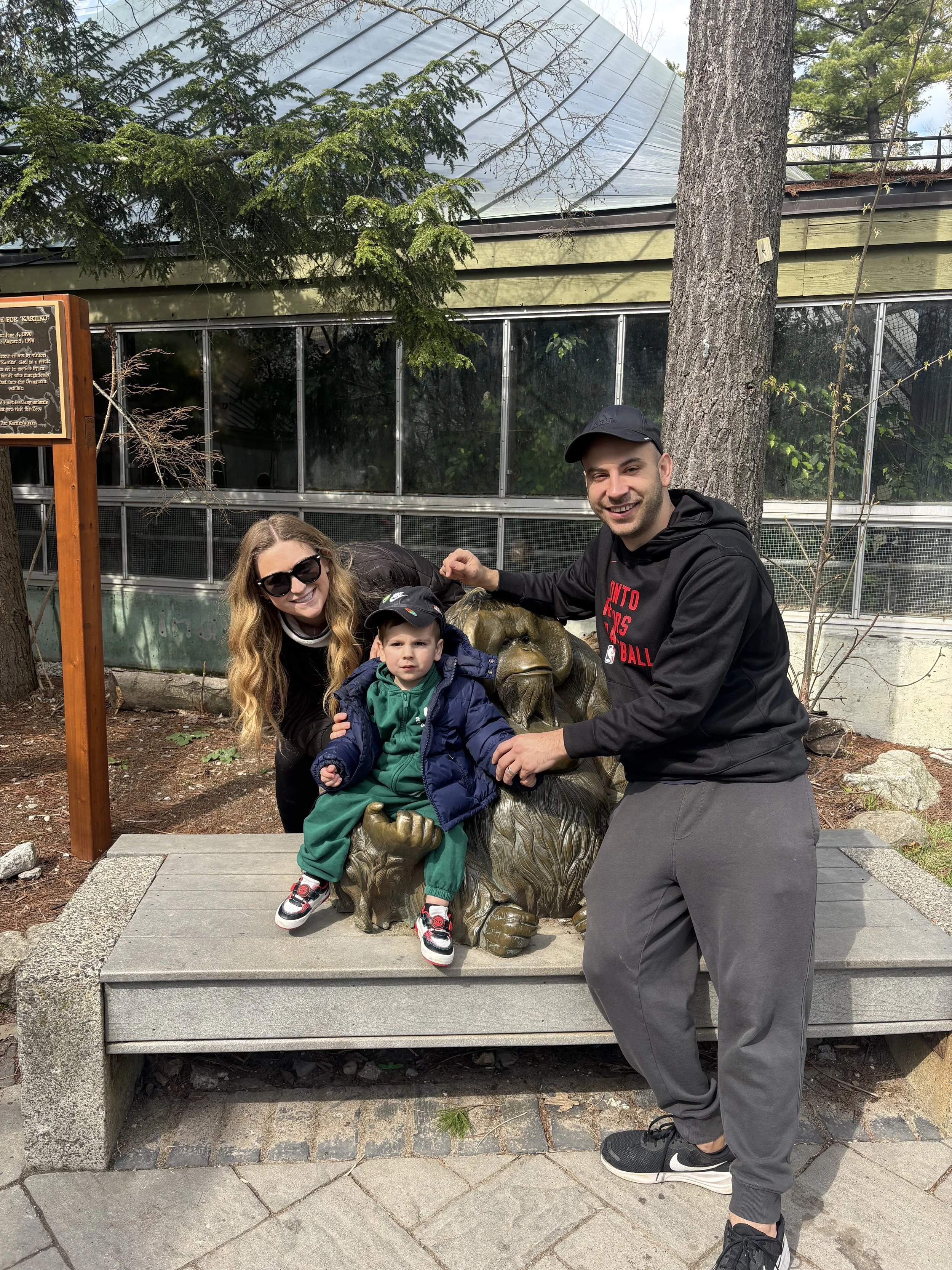 A family of three, including a woman, a man, and a young boy, posing together outdoors around a lion statue on a bench. The woman has long wavy blonde hair and is wearing sunglasses, the man is wearing a black cap and hoodie, and the boy is dressed in a green jacket and sneakers. They are smiling and appear to be enjoying a visit to a zoo or park. There are trees and a glass building in the background.