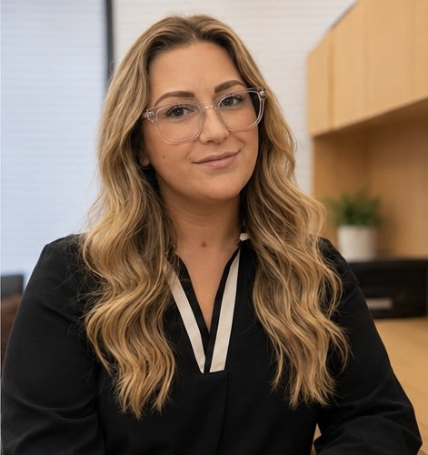 A woman with wavy blonde hair and glasses sitting in an office, wearing a black top with a striped collar.