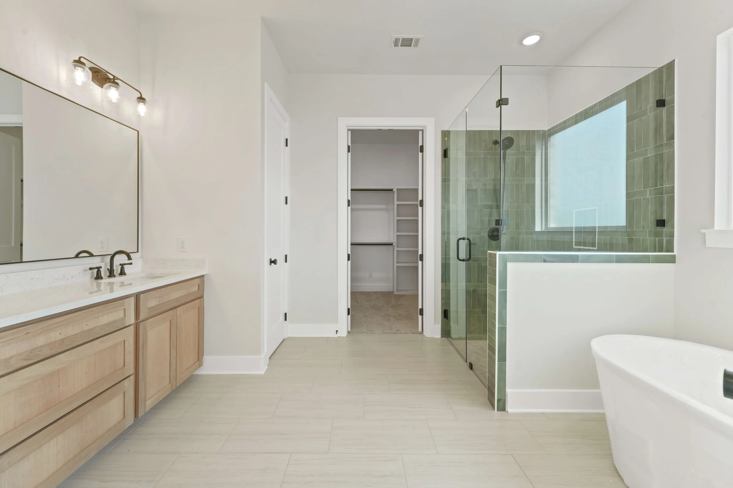 A modern bathroom with a wooden vanity, a large mirror, a walk-in shower with green tiles and glass doors, and a separate closet area visible through an open door.