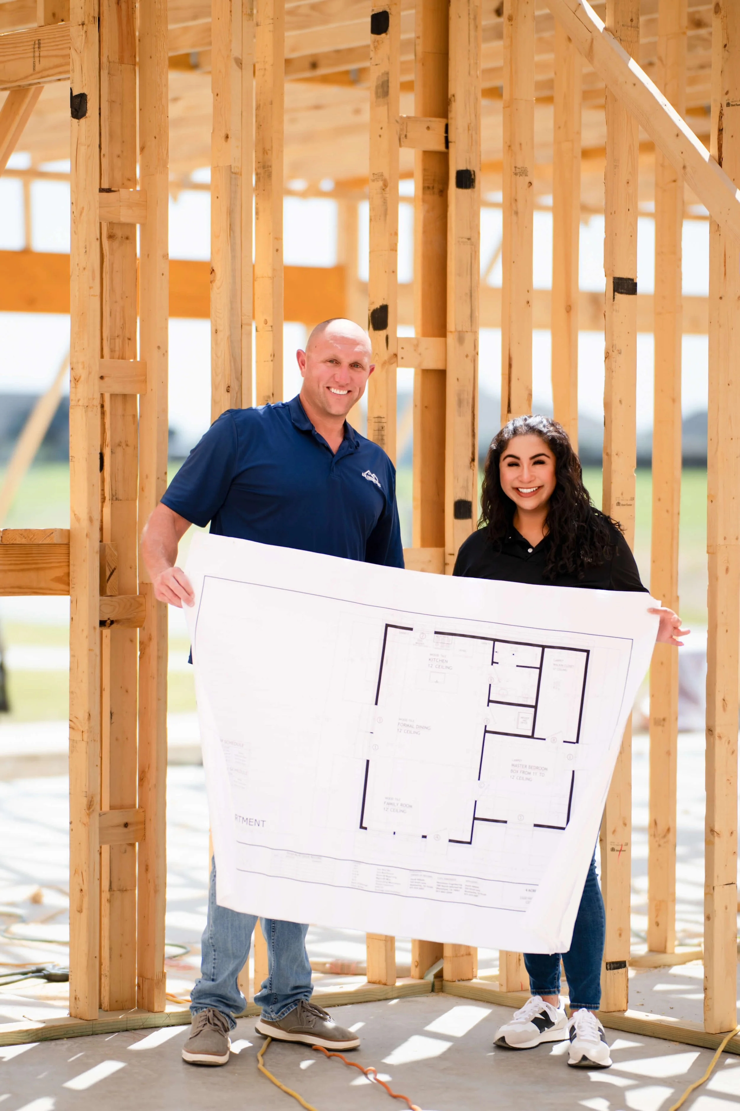 David and Tatum Abbott, owners of Azul Custom Homes, standing inside a wooden house frame on a construction site. They hold a large blueprint or architectural plan. The background shows the wooden framework of the house under construction.