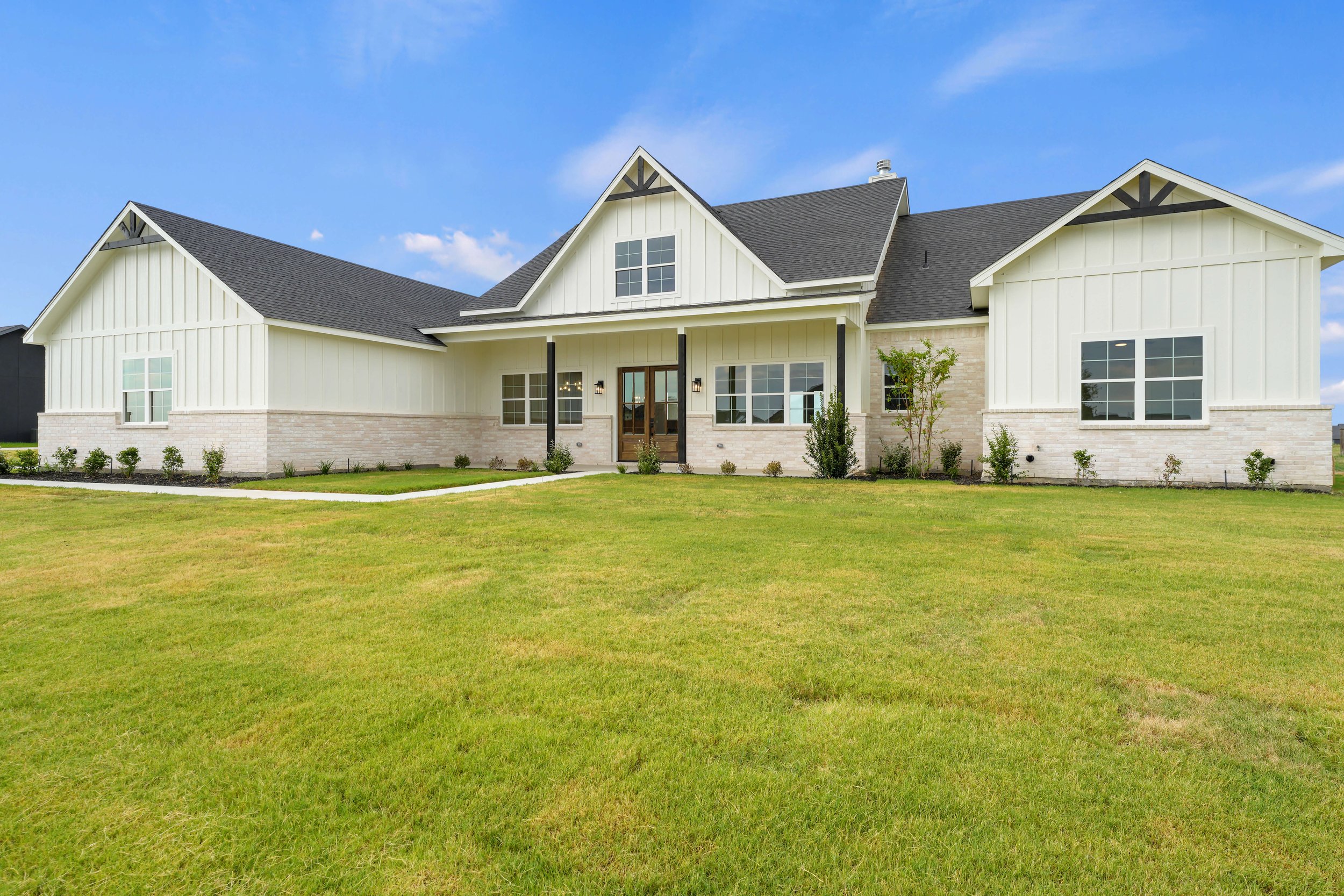 A modern white house with black accents, large windows, and a front yard with green grass and small bushes under a clear blue sky.