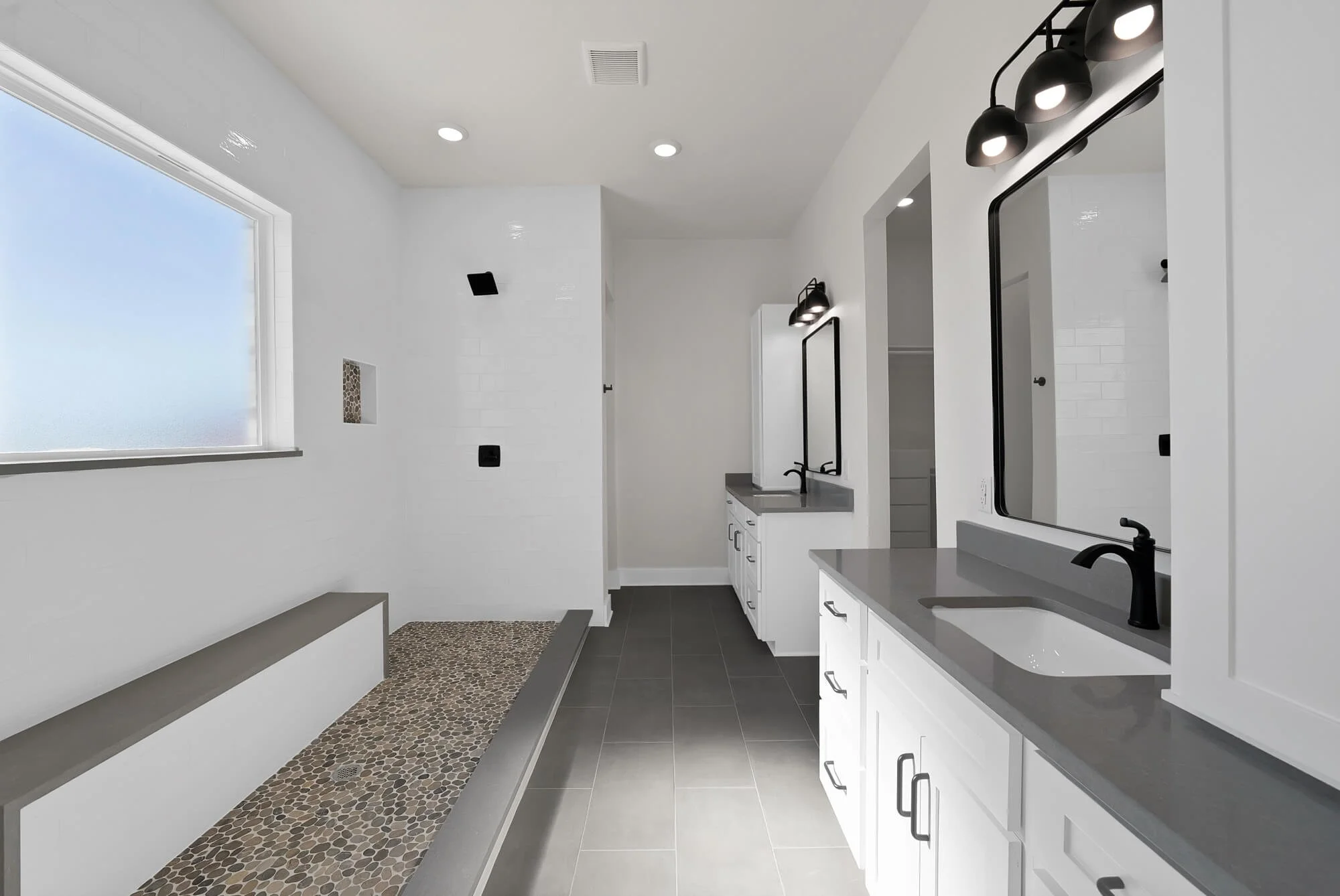 Modern laundry or utility room with white cabinets, a gray countertop, black fixtures, and a large frosted window, featuring a pebble stone floor and a black-framed mirror.