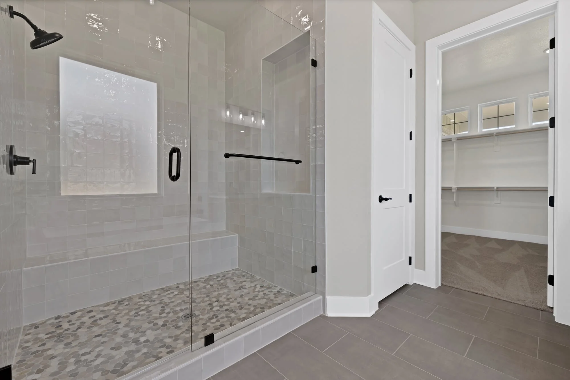 White tiled walk-in shower with a pebble floor, black fixtures, and glass doors, adjacent to a walk-in closet with white shelving.