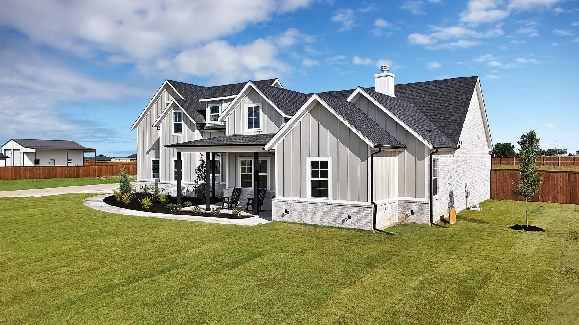 A modern two-story house with white exterior siding, a dark shingle roof, a chimney, and a covered front porch with benches. The house is surrounded by a well-maintained green lawn, a wooden fence, and newly planted trees and shrubs under a partly cl