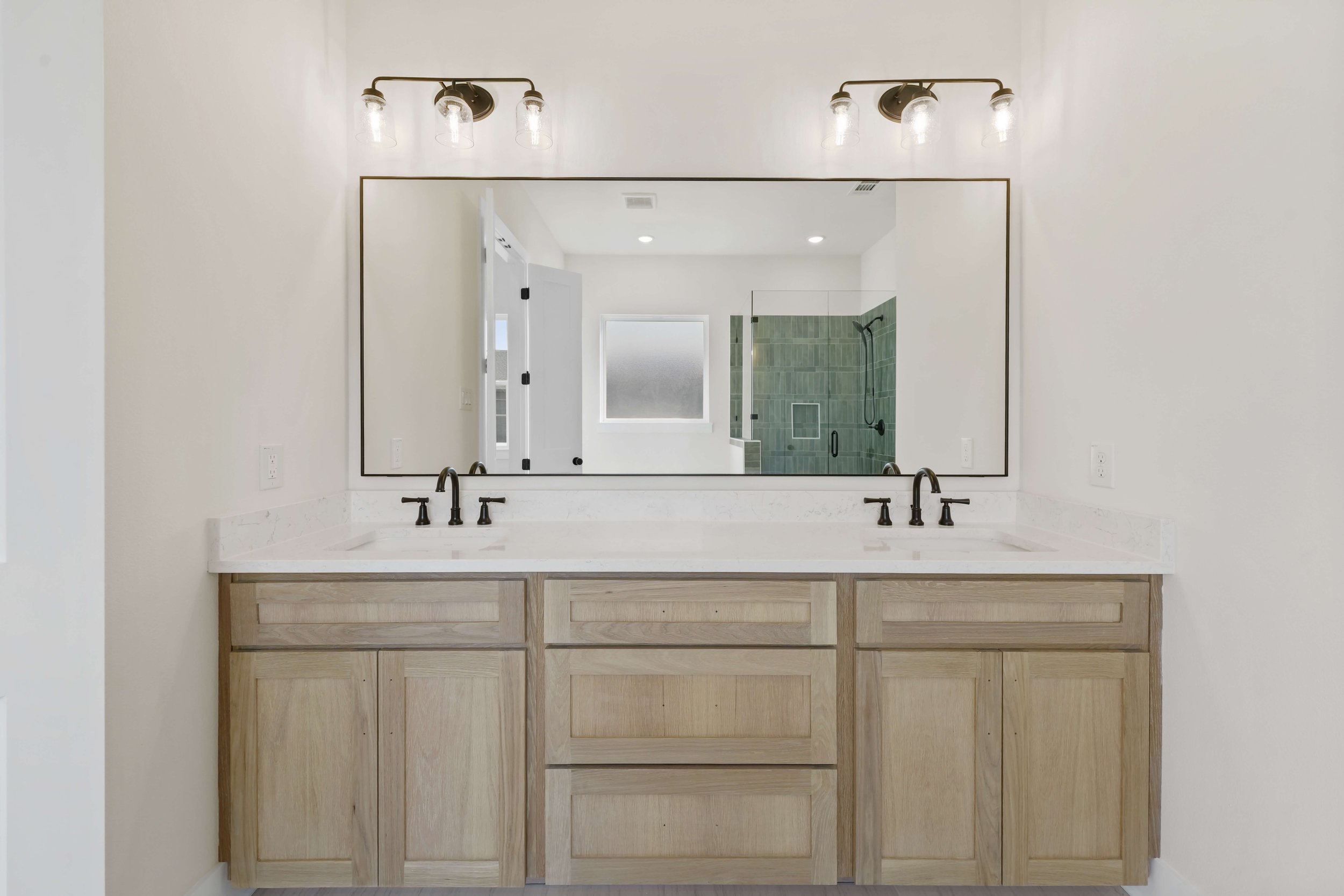 Modern bathroom vanity with a large mirror, black faucets, a white countertop, and a wooden cabinet base. Visible in the reflection is a shower with green tiles, a small frosted window, and a white door leading to another room.