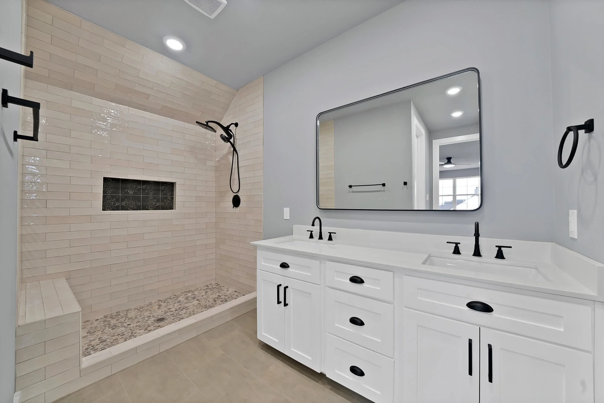 Modern bathroom with a walk-in shower featuring beige tiles, a black showerhead, and a built-in niche with decorative tiles. There is a white vanity with black hardware, a large mirror, and black fixtures, including a faucet and towel rings. The bath