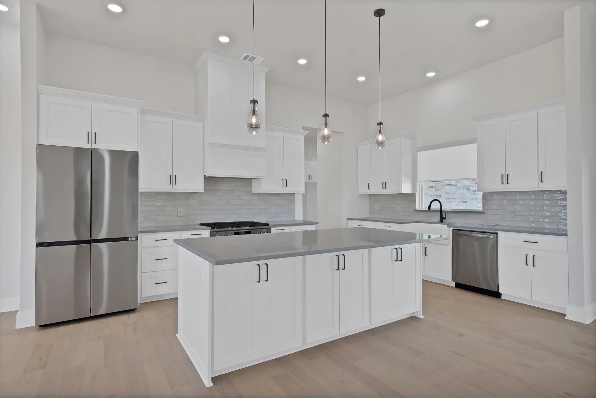 Modern white kitchen with stainless steel refrigerator, black handles, pendant lights, white cabinetry, gray countertops, and a light wood floor.