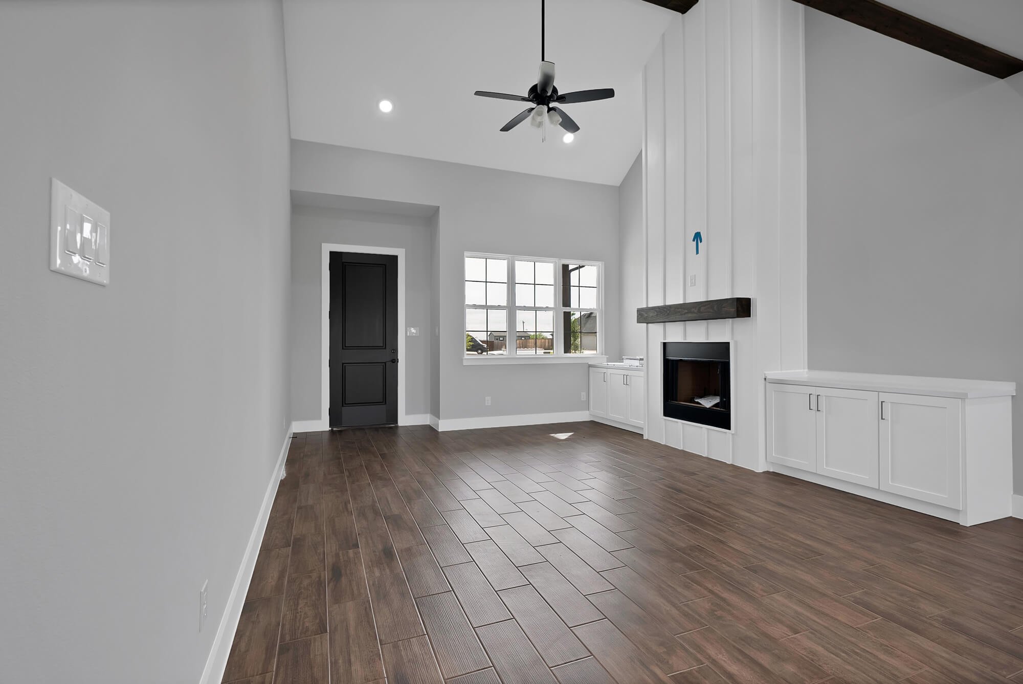 Interior of a modern, minimalist living room with white walls, dark wooden floor, a black door, two ceiling lights, a black ceiling fan, a window, and built-in white cabinets with a fireplace.