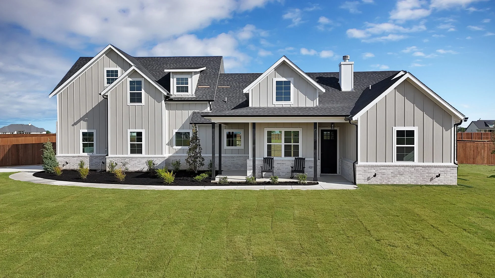 A modern gray house with white trim, a gabled roof, and multiple windows. The house has a front porch with two black chairs, a landscaped yard with grass and small bushes, and neighboring houses in the background under a blue sky with scattered cloud