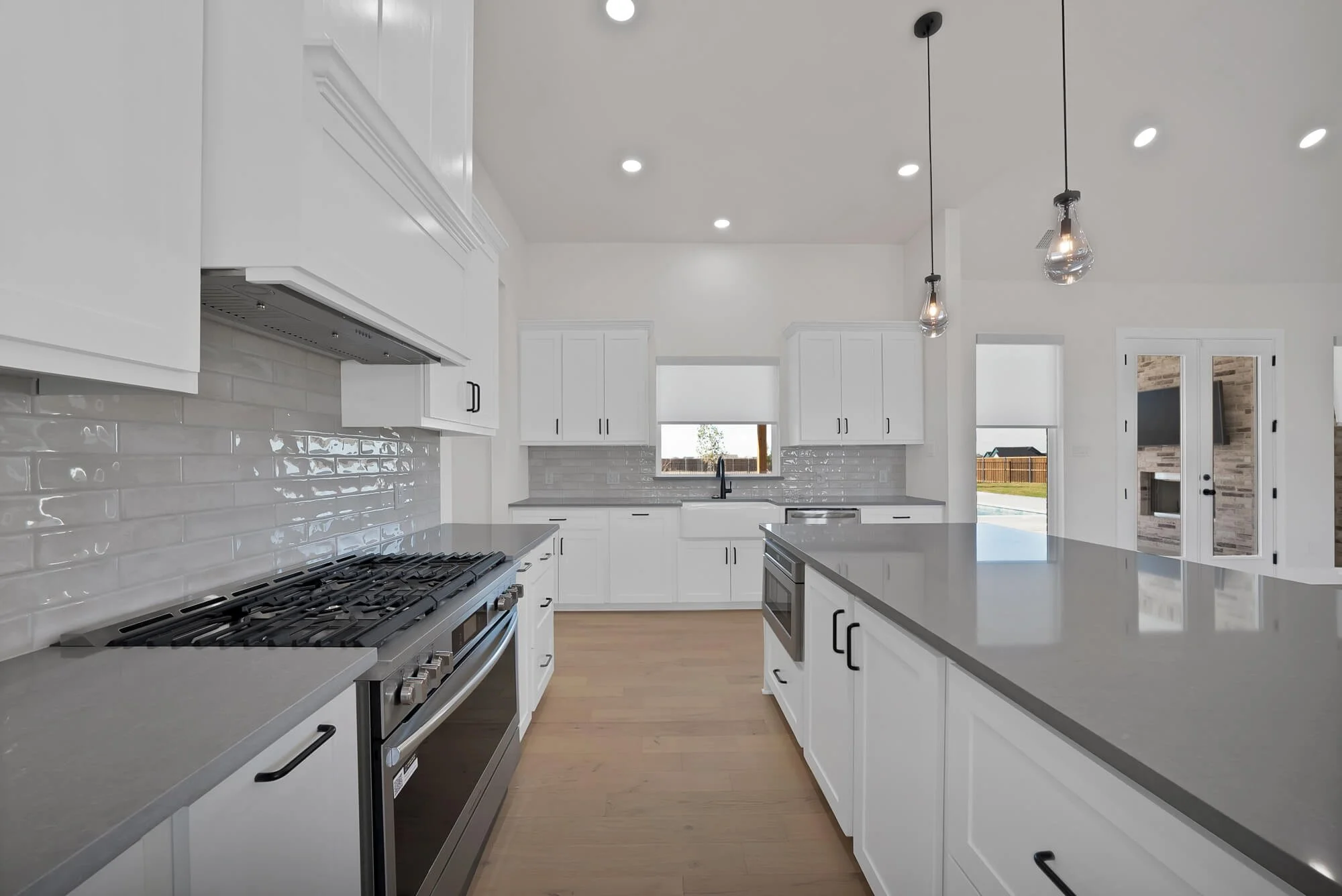 Modern white kitchen with stainless steel appliances, black handles, pendant lights, and large windows.