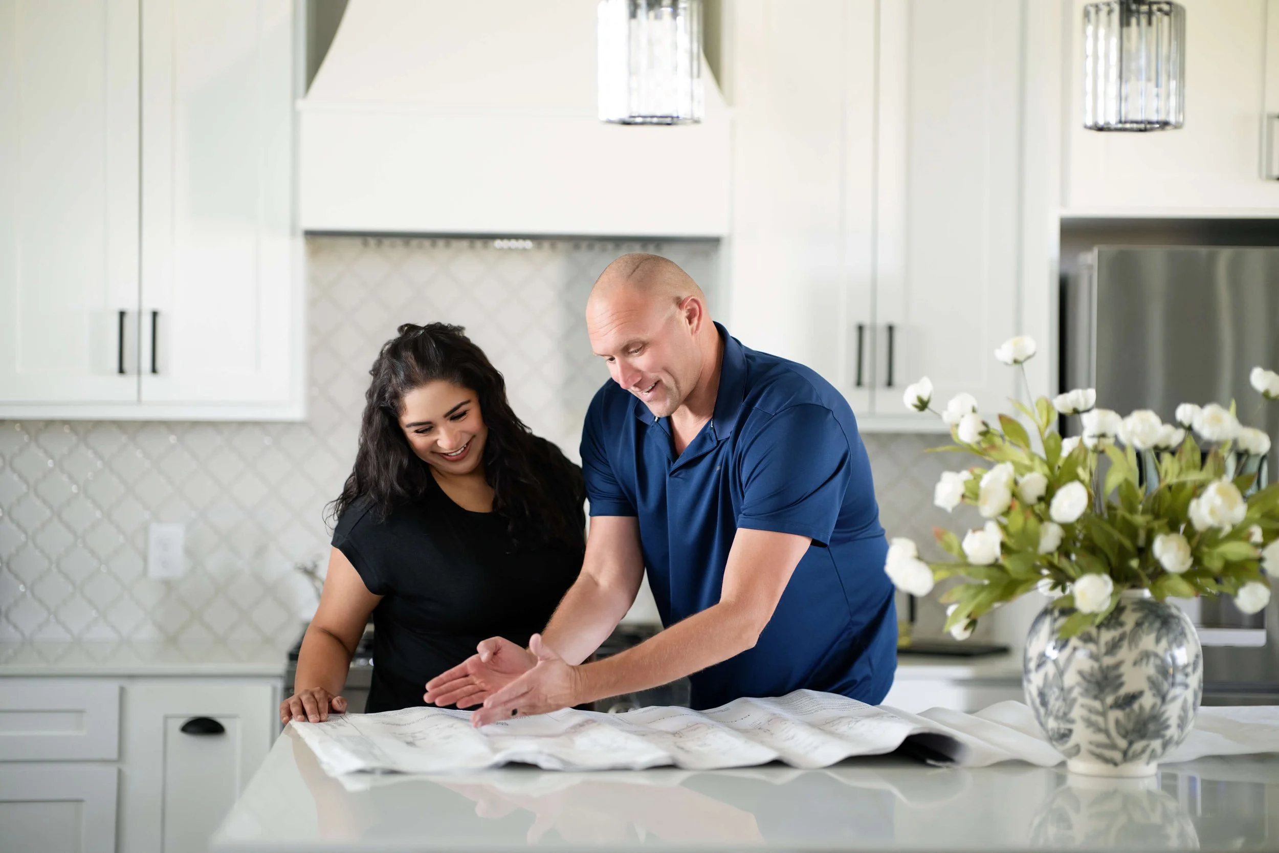 David and Tatum Abbott, owners of Azul Custom Homes, stand together in a modern kitchen, looking at a large sheet of paper or blueprint on the counter as they smile and discuss.