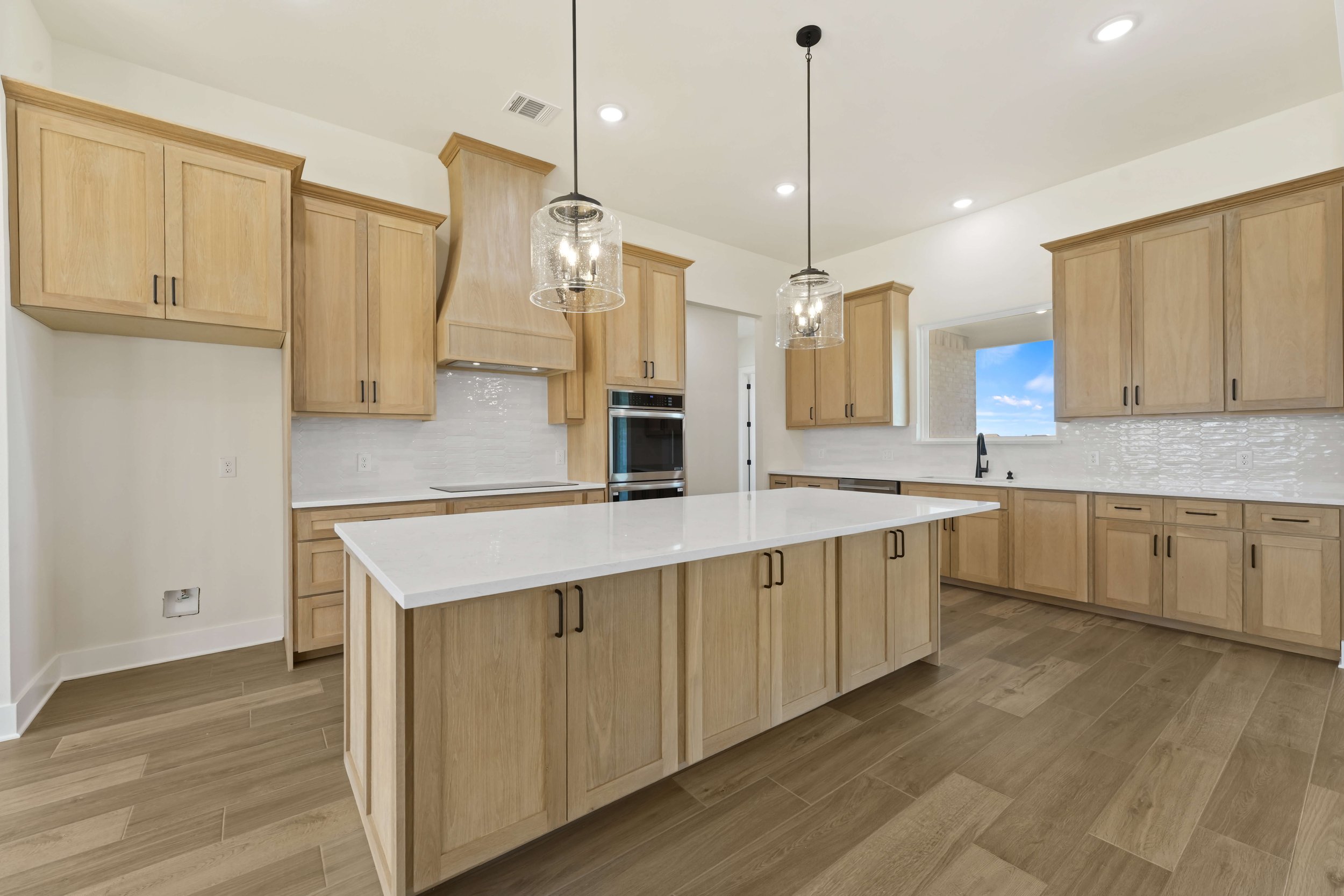 Modern kitchen with light wooden cabinets, white countertops, and a large kitchen island. Pendant lights hang over the island, and there is a window with a view of a blue sky and clouds.