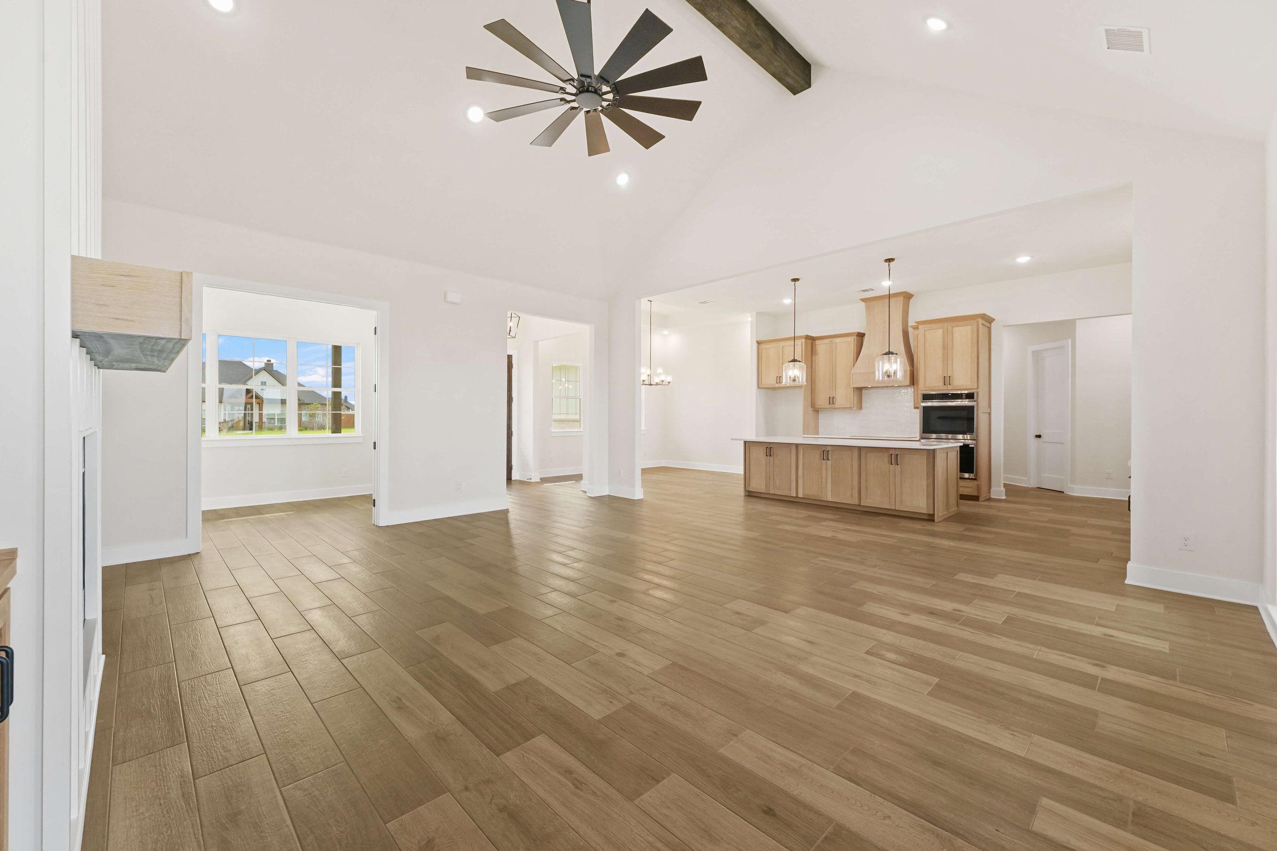 Empty open-concept living room and kitchen area with wood flooring, white walls, ceiling fan, large window, and natural light.