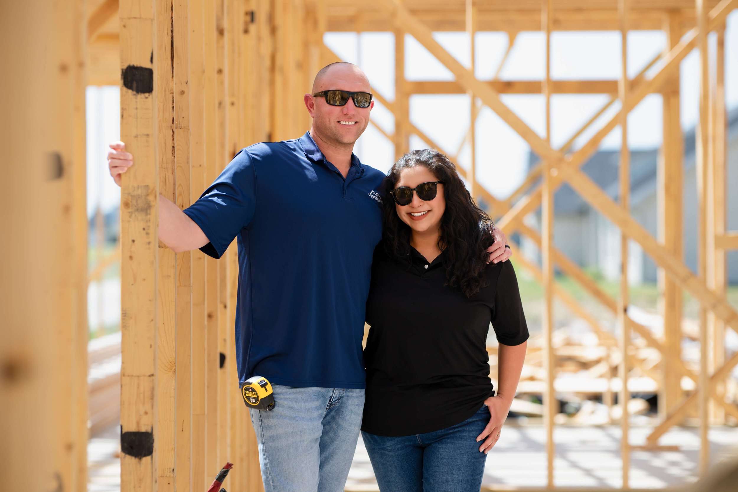 A man and woman wearing sunglasses at a building under construction with wooden framing. The man has a measuring tape attached to his jeans and is wearing a blue shirt, while the woman has a black shirt and dark hair.
