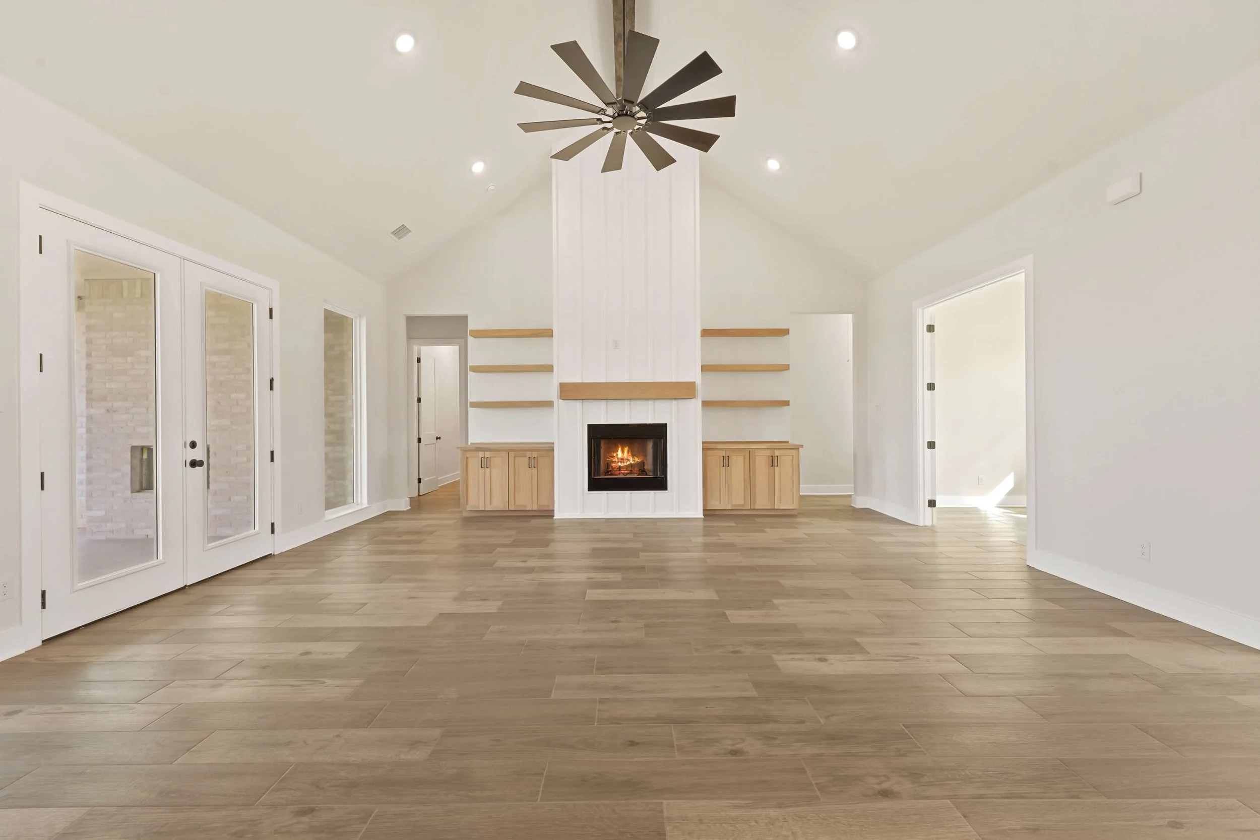 Living room with vaulted ceiling, white walls, a gas fireplace, built-in wooden cabinets, and built-in open shelves above the fireplace. Light wooden flooring and a large ceiling fan with black blades.