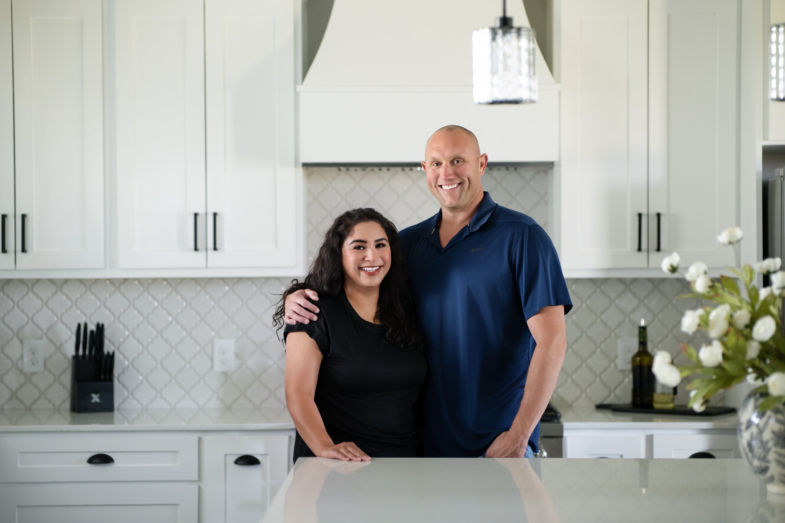 A couple stands in a modern kitchen, smiling at the camera. The woman has dark, curly hair and is wearing a black shirt, while the man is bald, wearing a blue polo shirt. White cabinets, a hood vent, and a tiled backsplash are visible in the background.