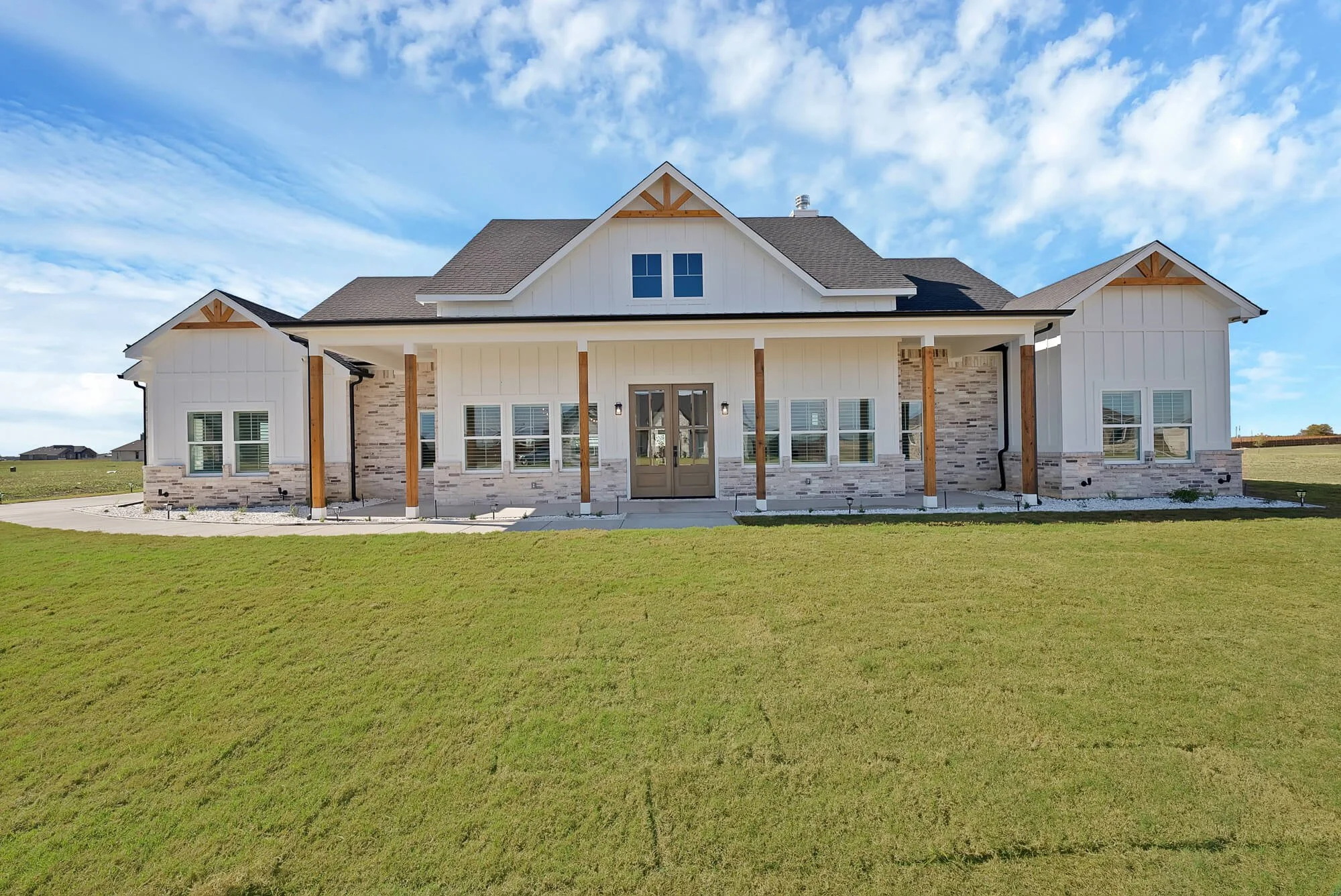 Front view of a modern house with white siding, stone accents, and a dark shingle roof, surrounded by a green lawn under a partly cloudy sky.