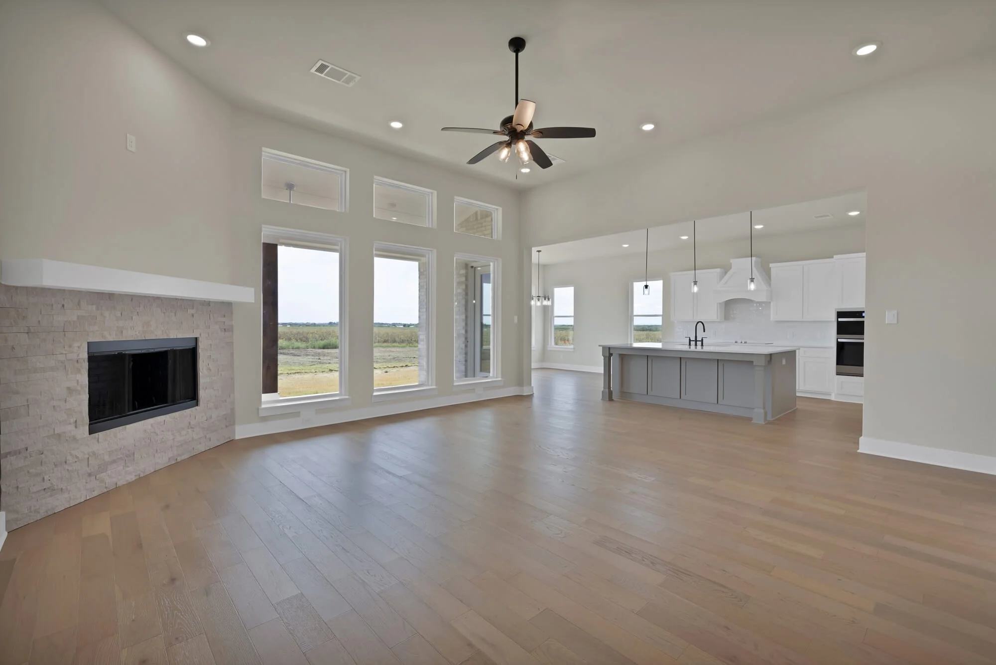 Open-concept living room and kitchen with large windows, hardwood floors, a ceiling fan, a fireplace with a brick surround, and white cabinetry in the kitchen.