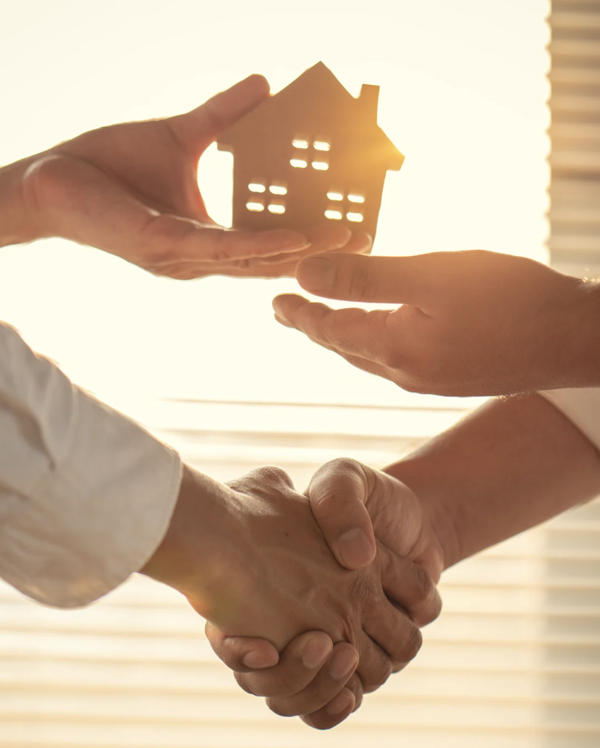 Two people shaking hands with sunlight shining through a window, one hand holding a small wooden house model.