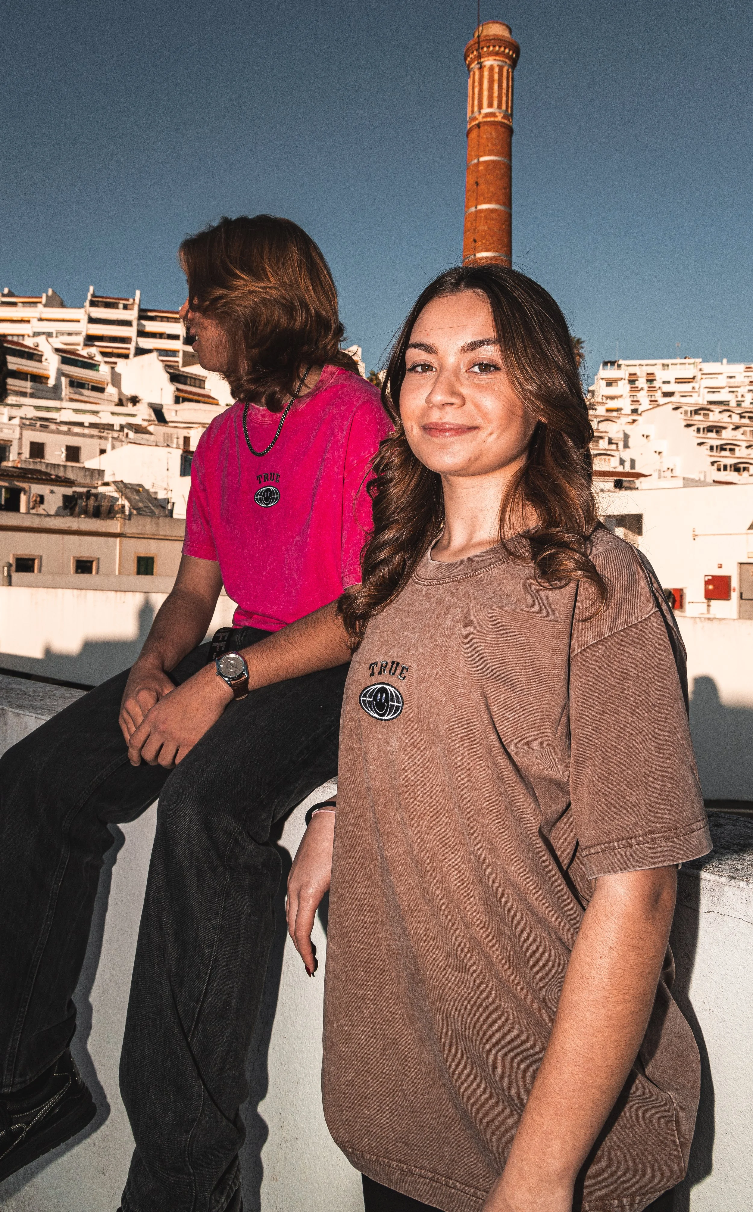Duas jovens mulheres com cabelo castanho, uma com cabelo liso e a outra com cabelo ondulado, posando em um terraço ao pôr do sol, com edifícios brancos e uma chaminé de tijolos ao fundo, vestindo camisetas com o logotipo 'TRUE'.