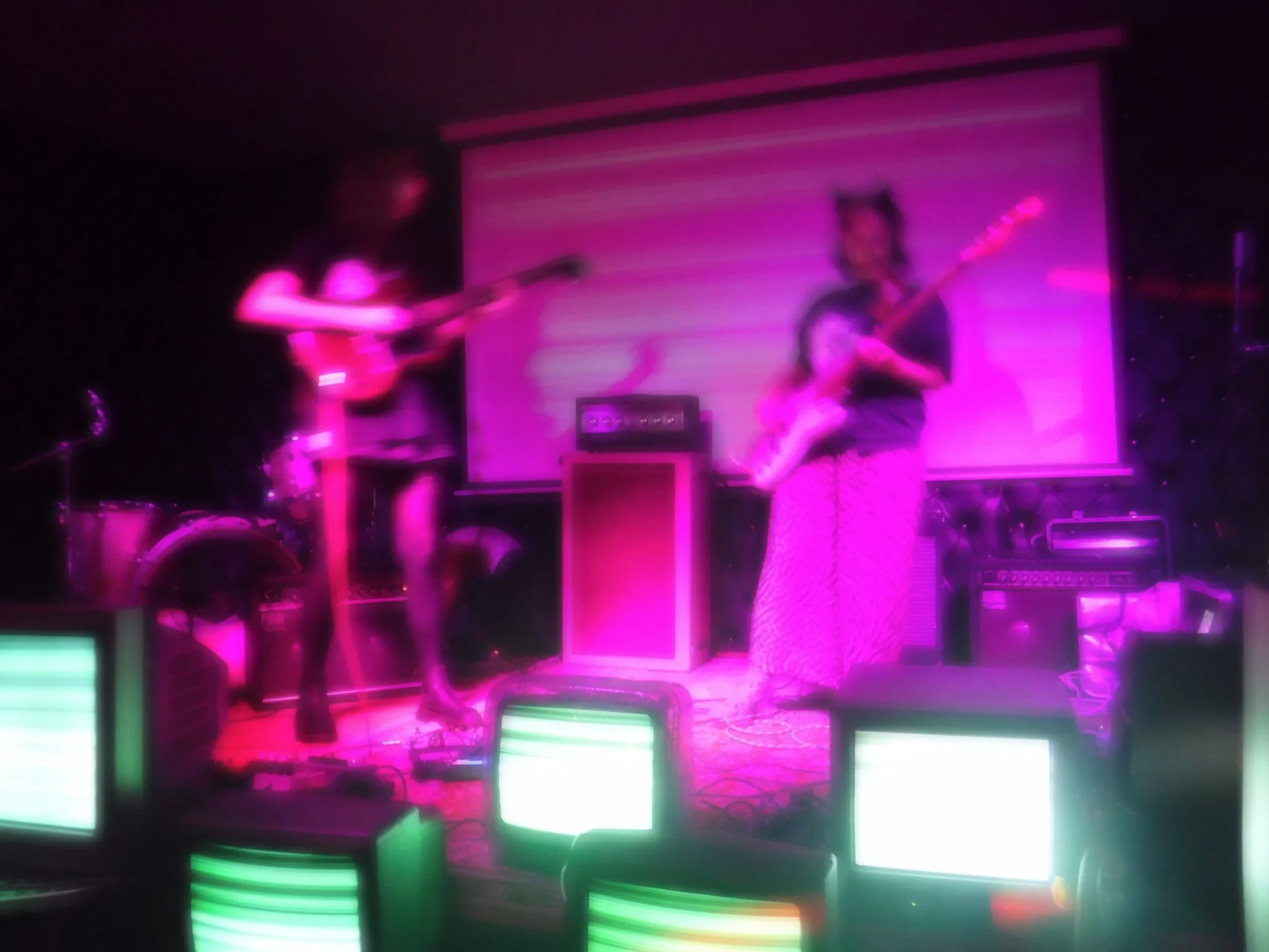Two people playing electric guitars on stage with pink and purple lighting, a drum set and amplifier in background, and a screen or backdrop behind them.