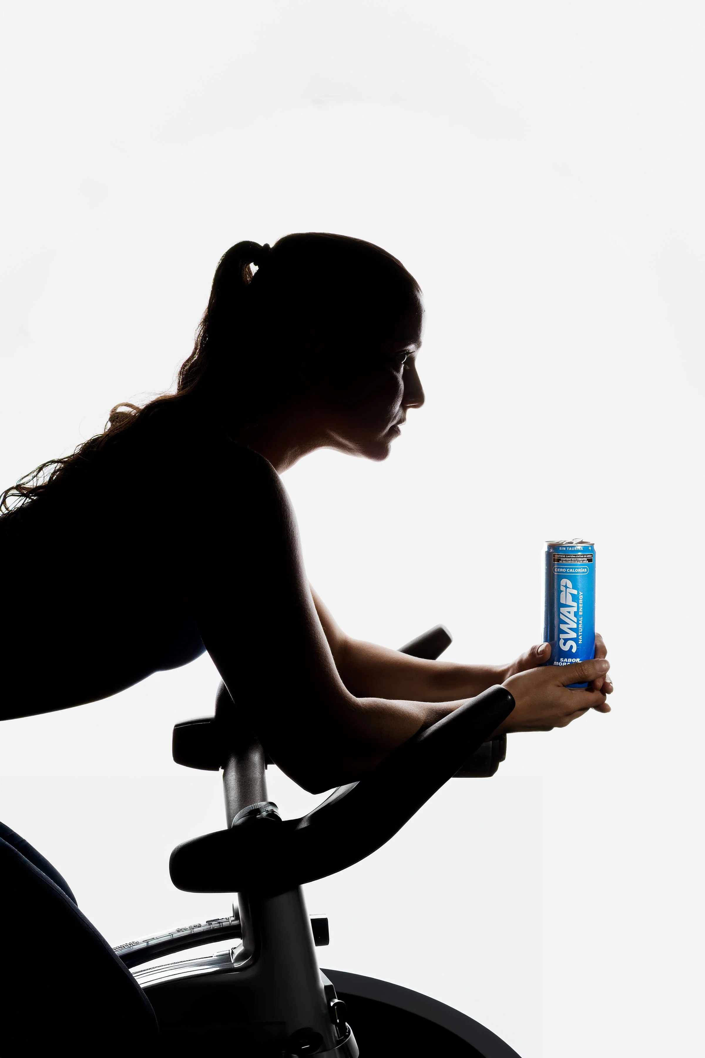 Silhouette of a woman with earbuds, sitting on a stationary bike, holding a can of energy drink, against a bright white background.