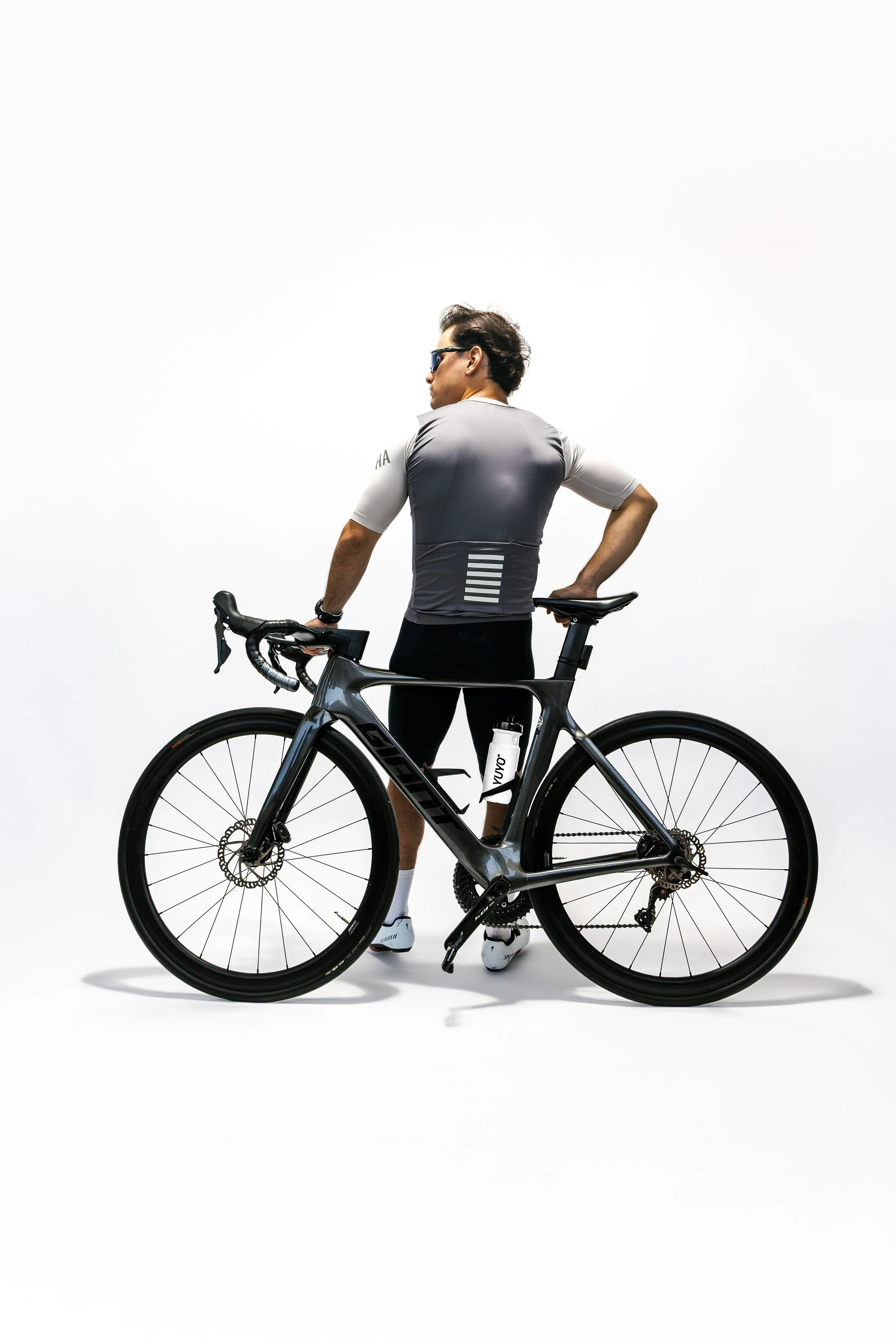 A man wearing sunglasses and cycling attire, standing next to a black road bicycle with a white water bottle, against a plain white background.