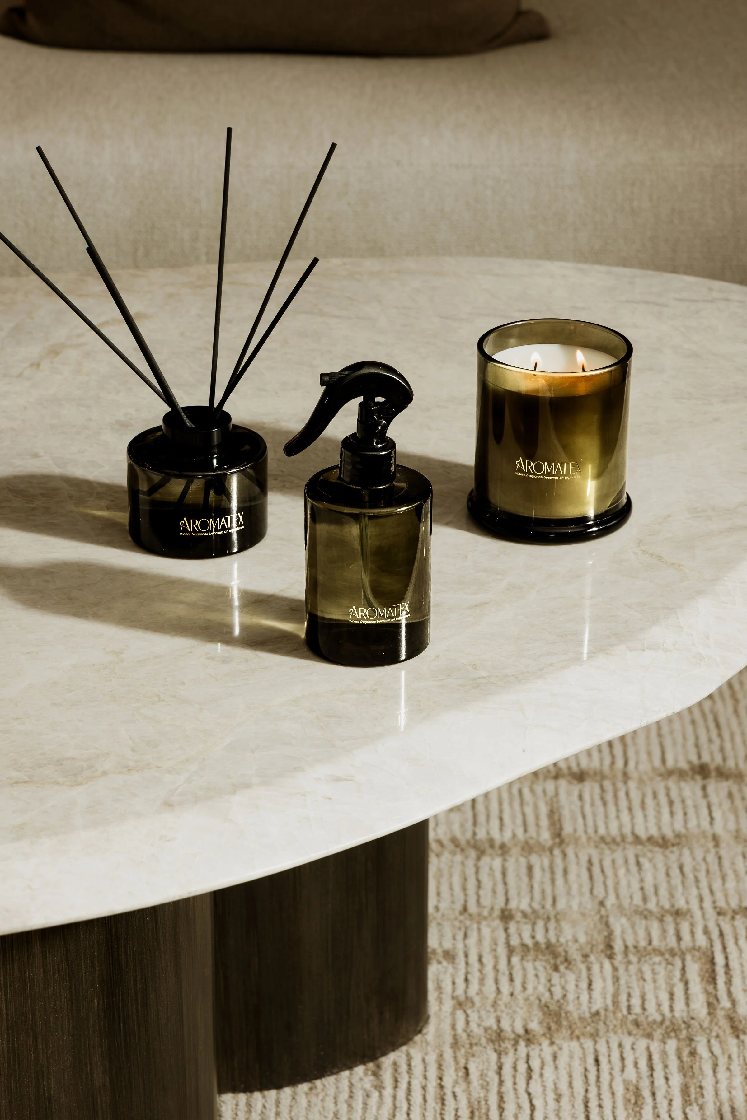 A black diffuser, a black spray bottle, and a candle in a black and gold holder on a white table. The diffuser has black sticks, and the candle is lit. The candle holder has the brand name 'AROMATEX' on it.