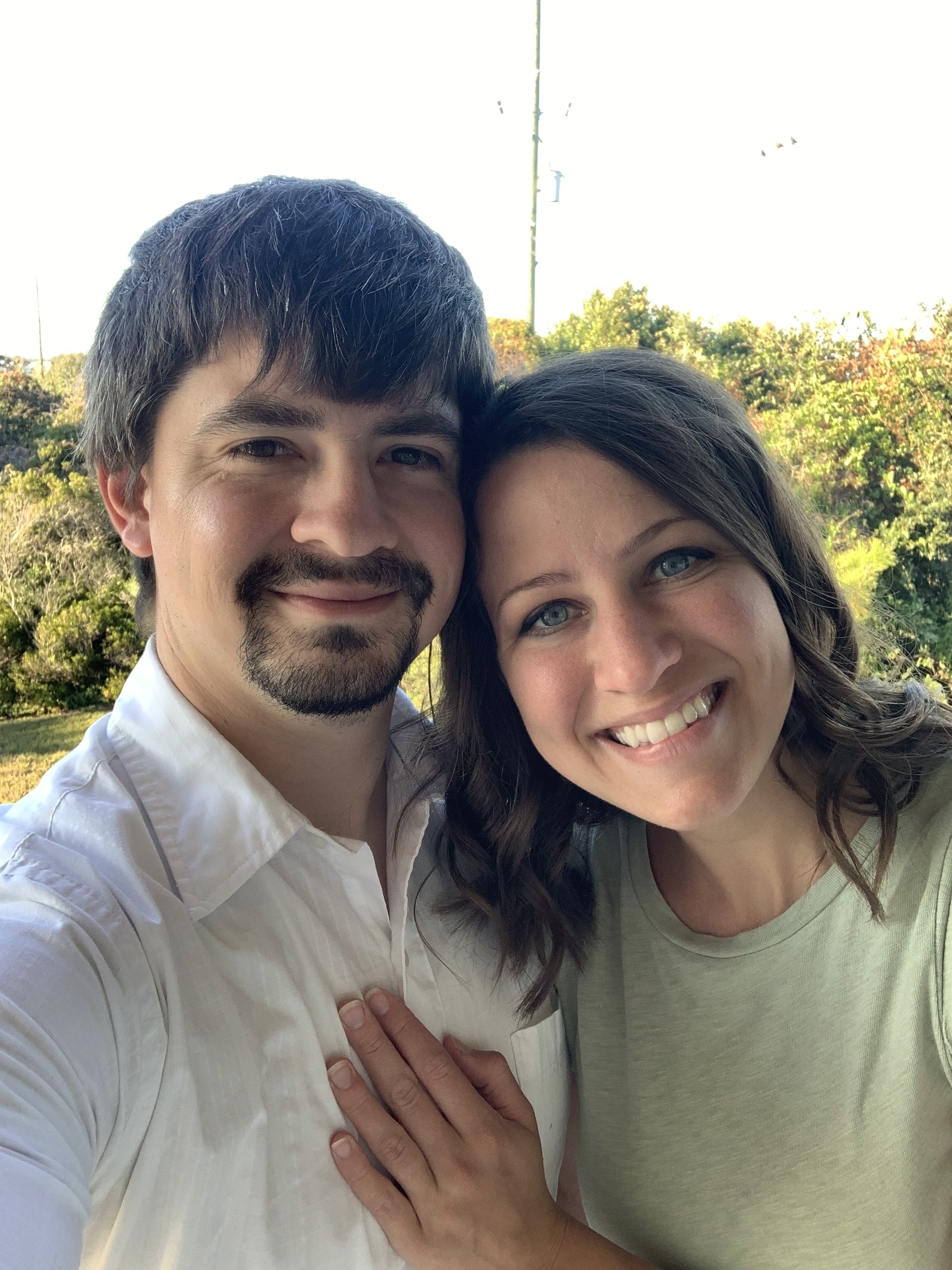 Close-up of a smiling man with dark hair and a mustache and beard, and a smiling woman with long curly hair, outdoors with greenery and trees in the background.