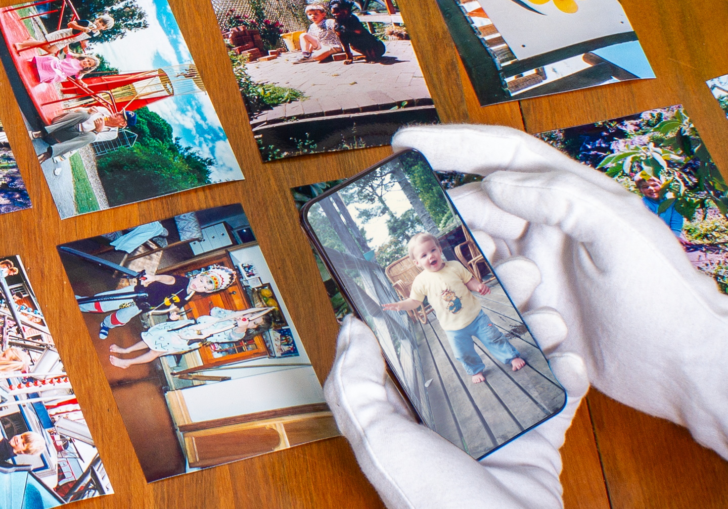 A person wearing white gloves is holding a smartphone, taking a picture of a young child standing on a wooden deck outside. The child is wearing a yellow shirt and blue jeans, and appears to be excited or surprised. Various printed photos are spread out on a wooden table, showing different scenes including children playing outdoors, people dressed in costumes indoors, and children on swings.