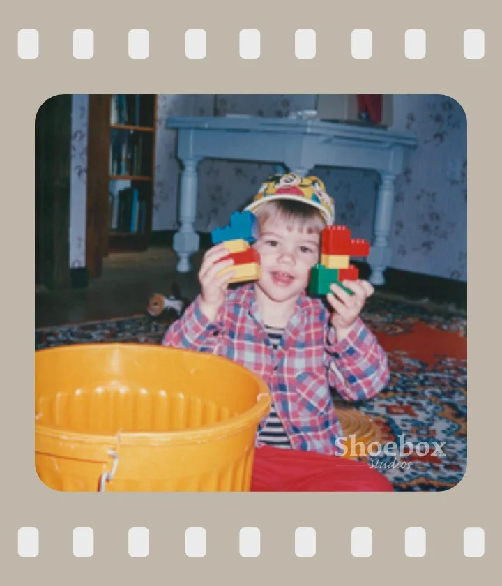 Young boy wearing a crown and plaid shirt playing with large LEGO bricks inside a house.