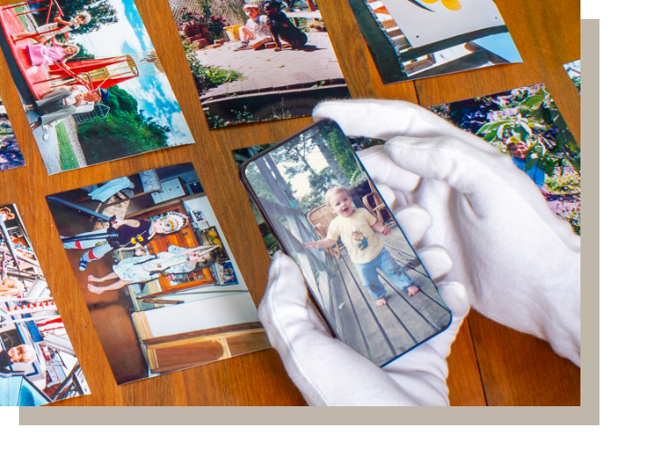 A person wearing white gloves is holding a smartphone and taking a photo of a young child with blonde hair, wearing a beige shirt and blue jeans, standing on a wooden deck.