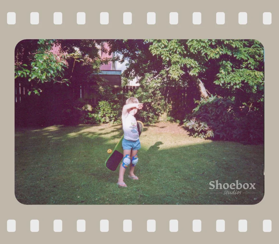 Child standing in a yard, holding skateboard, shielding their eyes from the sun, surrounded by green trees and bushes.