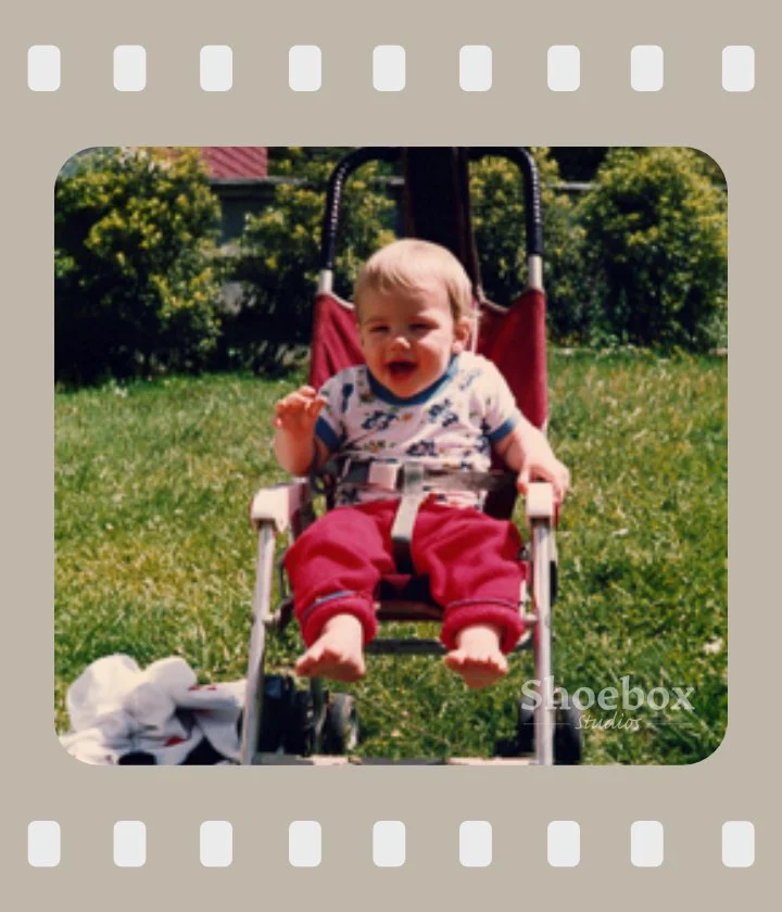 A young child sitting in a red stroller outdoors on a grassy lawn, smiling and waving, with bushes and a house in the background.