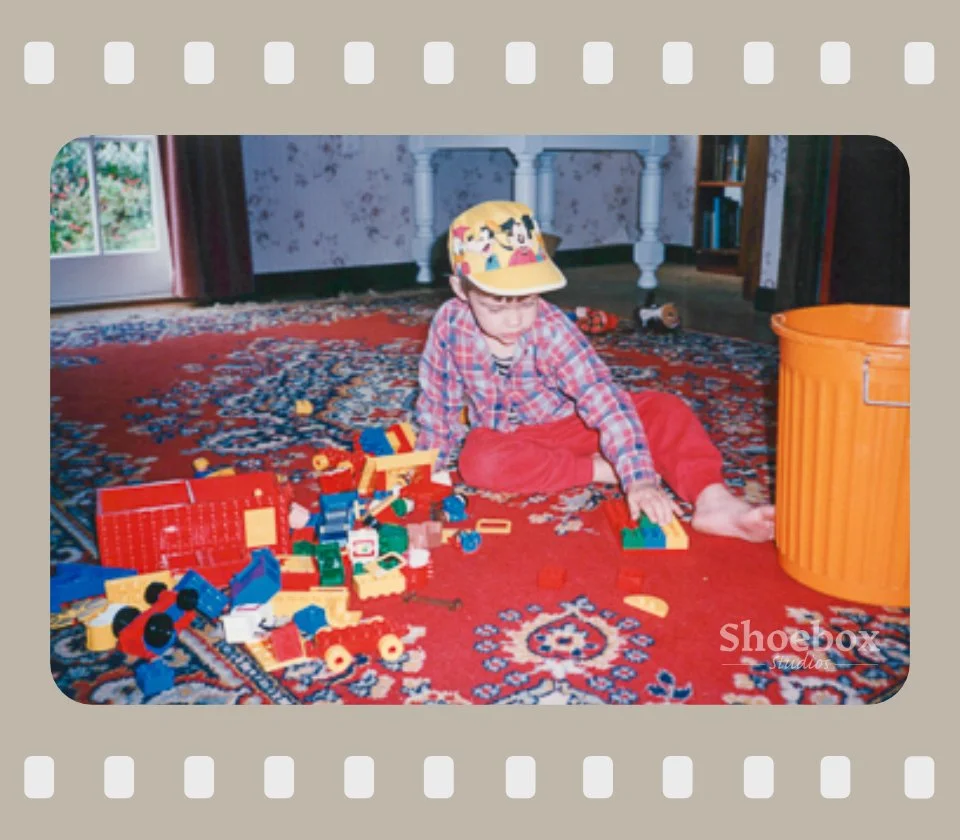 A young boy sitting on a red patterned carpet, playing with colorful building blocks next to a large orange trash can inside a house.