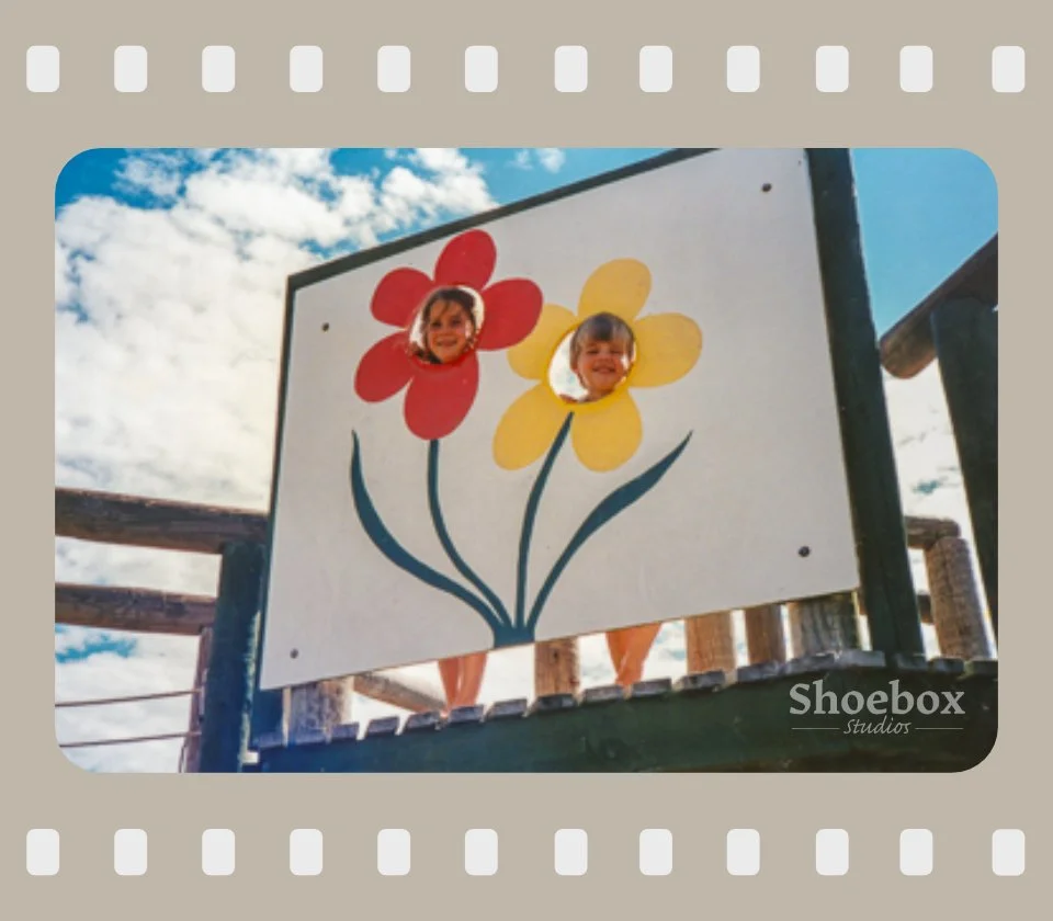 Two children playing at a park, peeking through flower-shaped holes in a painted board with a red flower and yellow flower, under a blue sky with scattered clouds.