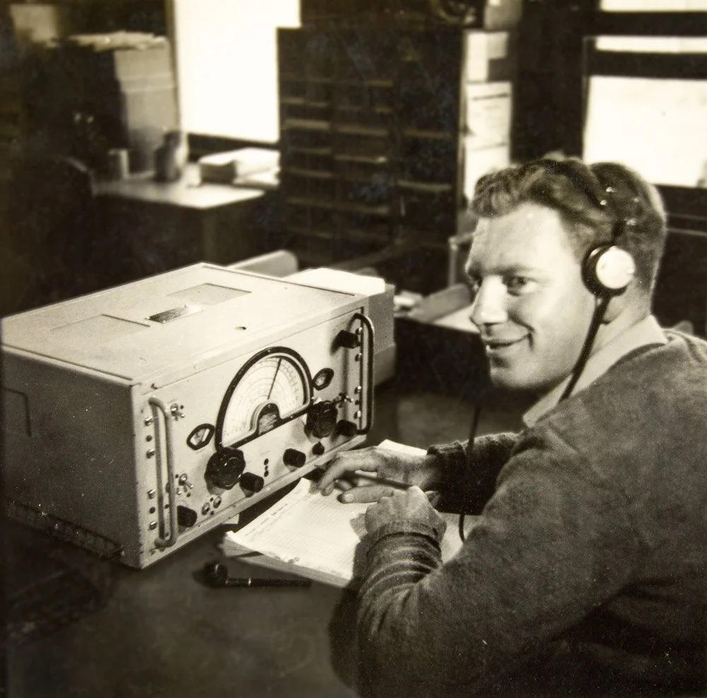 A man with headphones working at a vintage radio or communication device in an office setting, smiling while looking to the camera.
