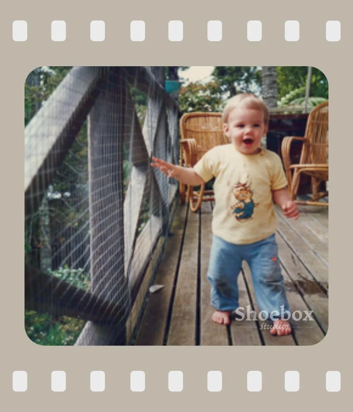 A young child standing on a wooden deck, touching a wire mesh fence with a tropical outdoor setting in the background.