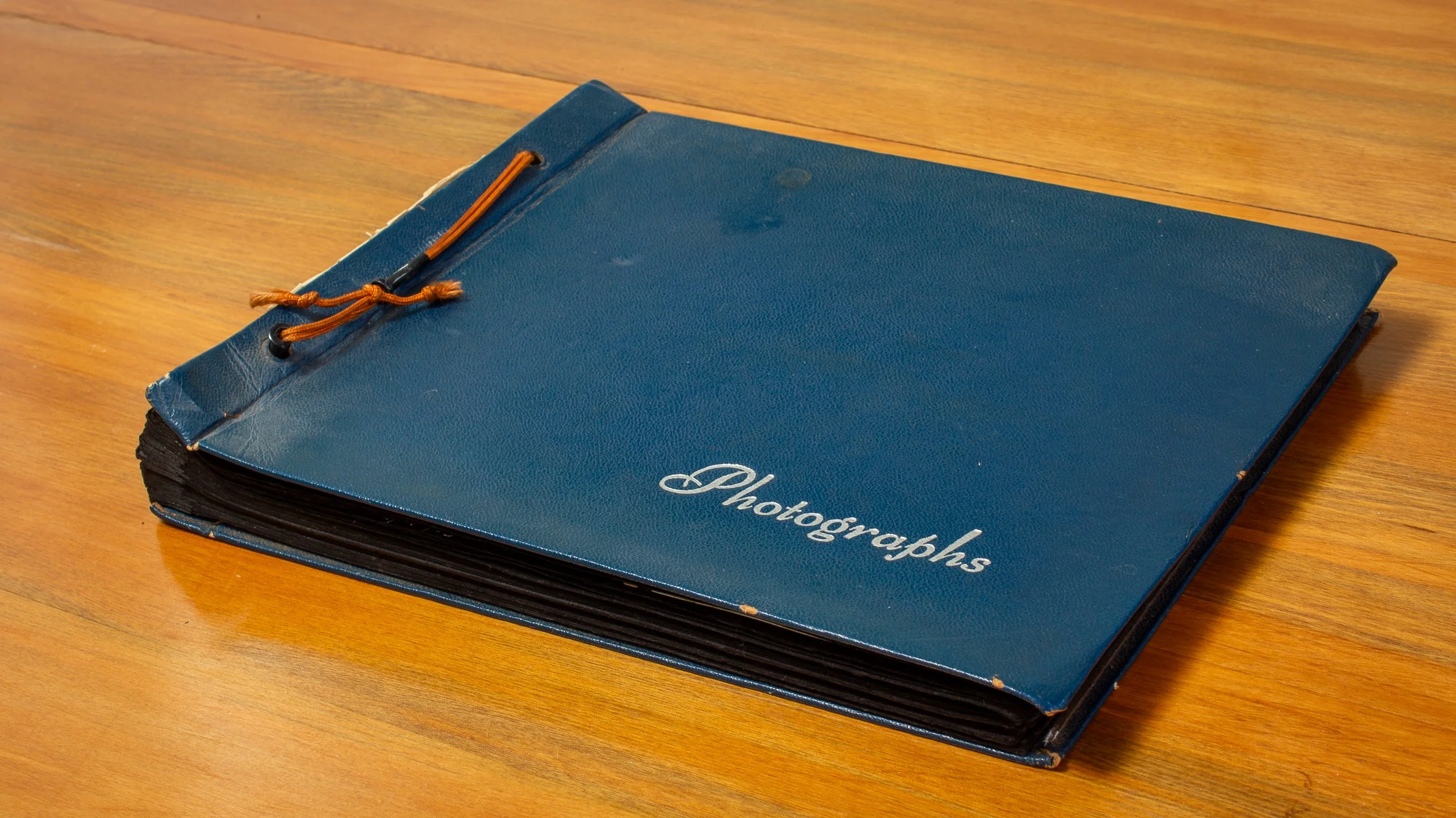 A blue hardcover photograph album with an orange elastic band and black pages, resting on a wooden surface.